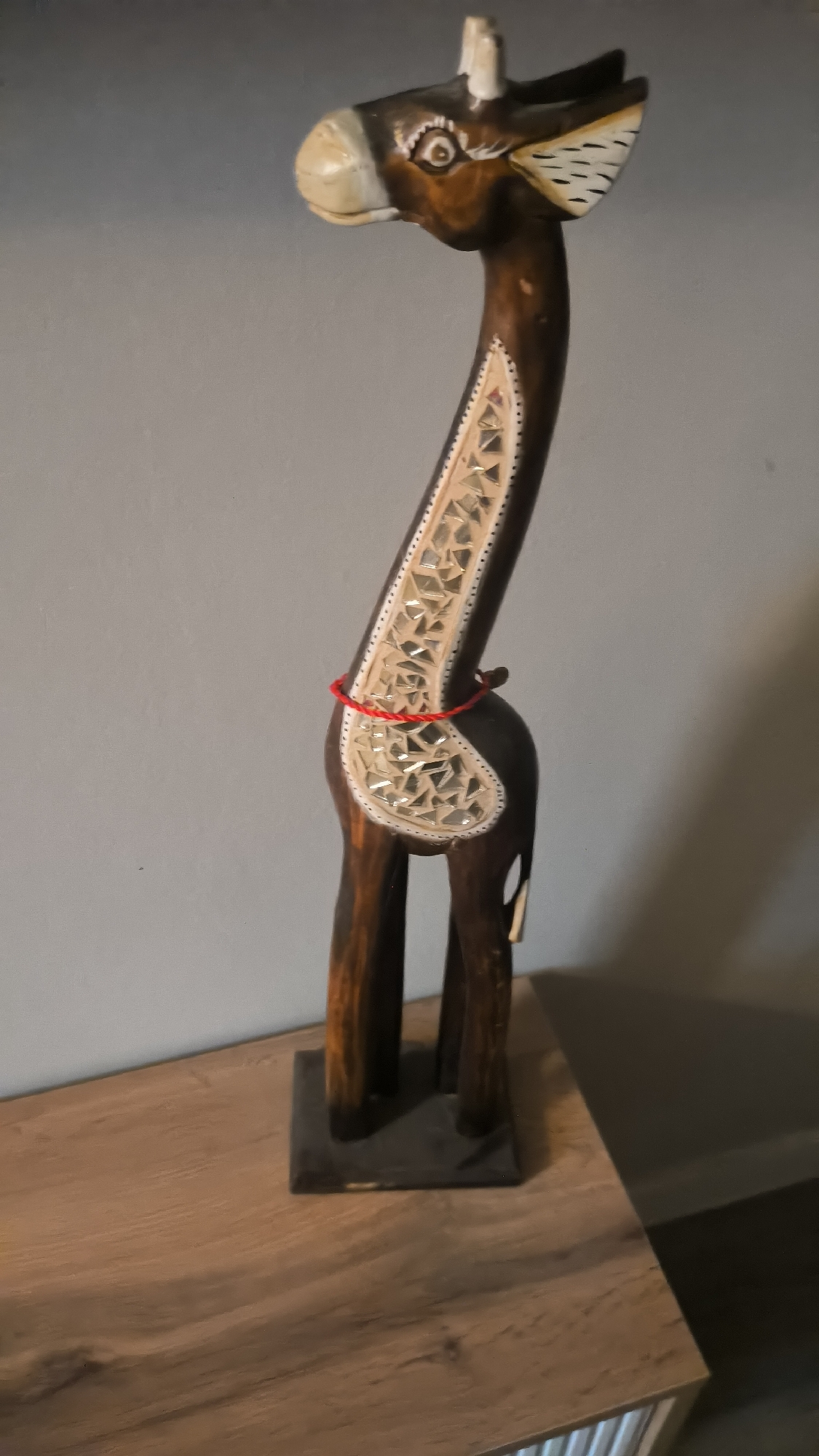 A wooden giraffe figurine, approximately 2 feet tall, stands on a wooden surface. The giraffe is dark brown with lighter accents on its face and a cut-out pattern on its neck filled with reflective material. A thin red string is tied around its neck. The figurine is positioned against a plain grey wall. The lighting suggests an indoor setting, possibly a home environment. No people are present. The location is not specified as Kovrov, Russia, nor is there any indication of a city or country.