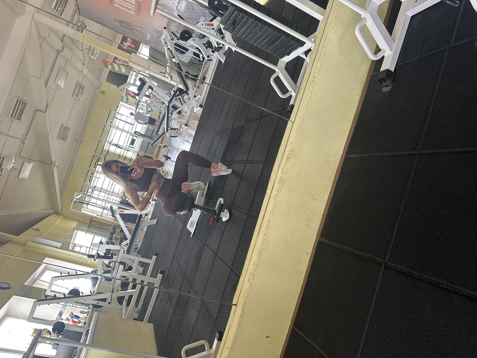 A woman takes a selfie in a mirror in a gym in Chaikovsky, Russia. The gym has a rubber floor and many weight machines.