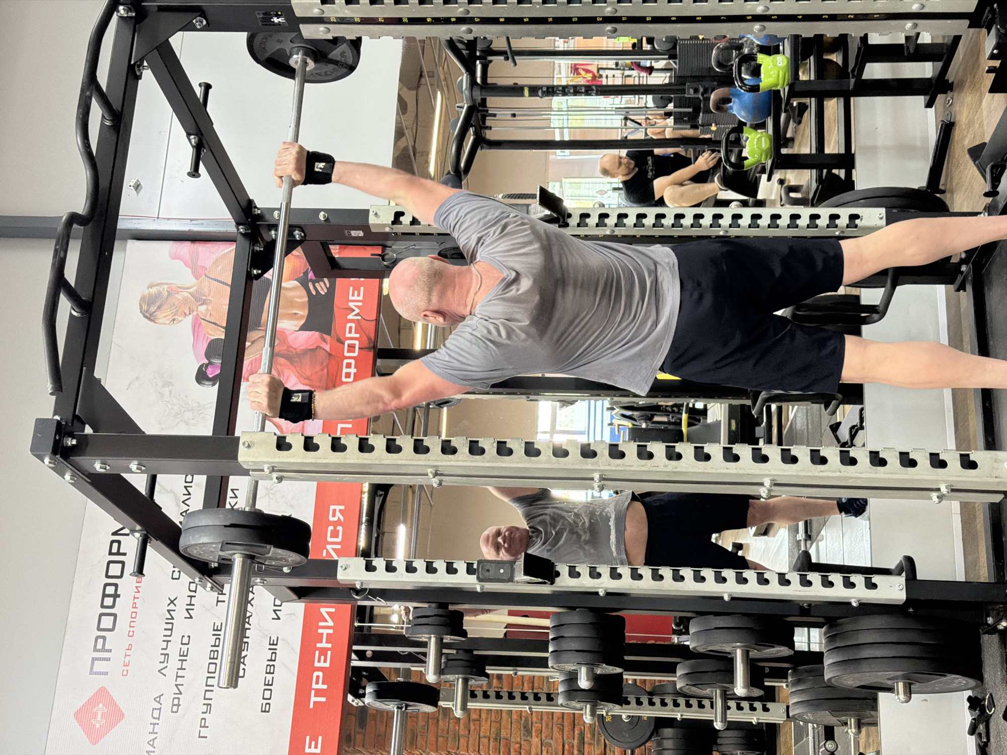 A man in a gray t-shirt and black shorts is performing a barbell exercise within a weight rack at a gym. He is holding the barbell with both hands, wearing wrist supports. The setting includes a mirror reflection showing the man and other gym-goers in the background. The location is Chaikovsky, Russia, as indicated by the Russian text "ПРОФОРМ" and other Cyrillic text on the equipment and wall posters in the image. Weight plates and other equipment are visible around the weight rack.