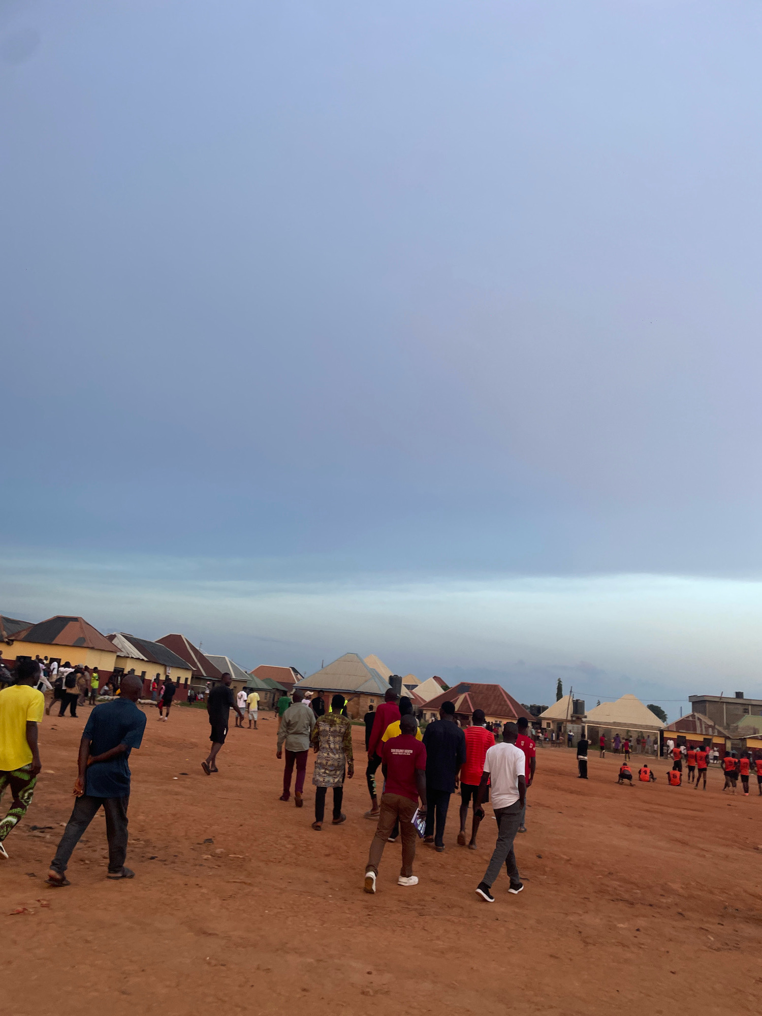 A group of people are gathered on a dirt road in a residential area of Kurunduma, Nigeria. The sky is overcast with pale blue and grey clouds. In the background, there are several buildings with corrugated metal roofs in various colors, including yellow, brown, and red. Some buildings appear to be homes, while others might be shops or community buildings. In the foreground and mid-ground, a diverse group of individuals, primarily men, are walking and standing. Some are dressed in casual attire, such as t-shirts and trousers, while others are wearing what appears to be sports uniforms, including orange vests and red shirts. There is a sense of activity and gathering, possibly related to an event or local activity. The ground is a reddish-brown dirt, indicative of the local terrain. Some of the people are interacting with each other, while others are walking in different directions. The overall impression is of a community gathering in a typical Nigerian urban or semi-urban setting.