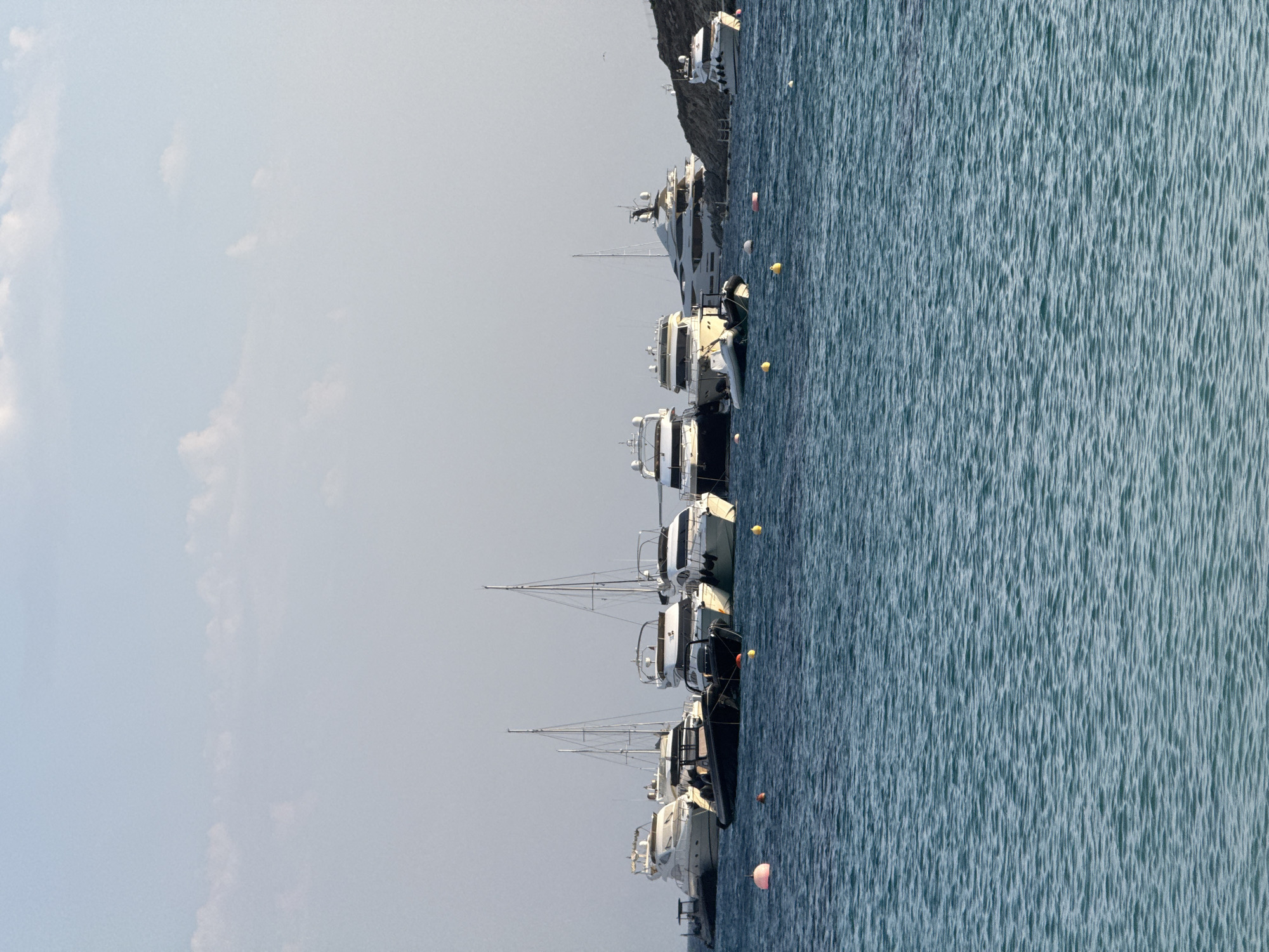 The image depicts a scenic view of a bay with several yachts and smaller boats moored in the water, situated in Agios Ioannis Diakoftis, Greece. The sea is a vibrant turquoise, with gentle ripples on its surface. In the background, a rocky shoreline with sparse vegetation is visible, meeting a hazy, pale blue sky with a few scattered clouds. The boats, varying in size and design, are docked in a relatively close proximity, creating a picturesque maritime scene. Among them are large, sleek white yachts with multiple decks and masts, suggesting luxury and leisure. Smaller, more utilitarian boats, including a dark-colored rigid inflatable boat, are also present. Several buoys, including a prominent pink one, are floating in the water, marking the anchorage area. The overall atmosphere is serene and tranquil, indicative of a calm day at sea.