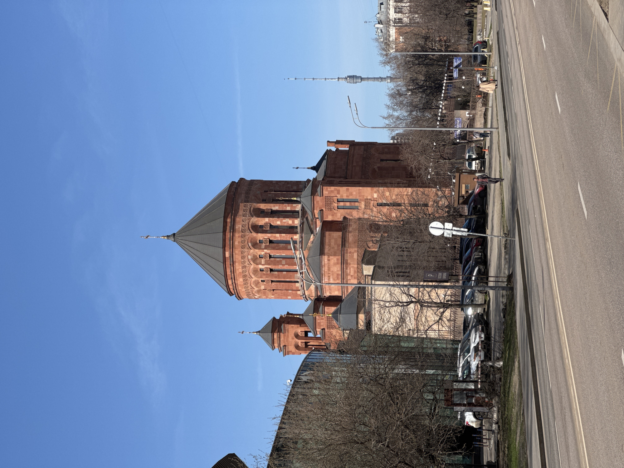 A low-angle, medium-distance shot depicts the Saint Gregory the Illuminator Cathedral in Yerevan, Armenia. The cathedral is made of reddish-brown stone with a conical gray roof, featuring rounded arches and vertical windows. In the background, a television tower rises into the clear blue sky. Alongside the building is a multi-lane road with parked cars and leafless trees. People are standing on the sidewalk. The environment appears to be urban. The geographical location of Khimki, Russia is not visible in this image.