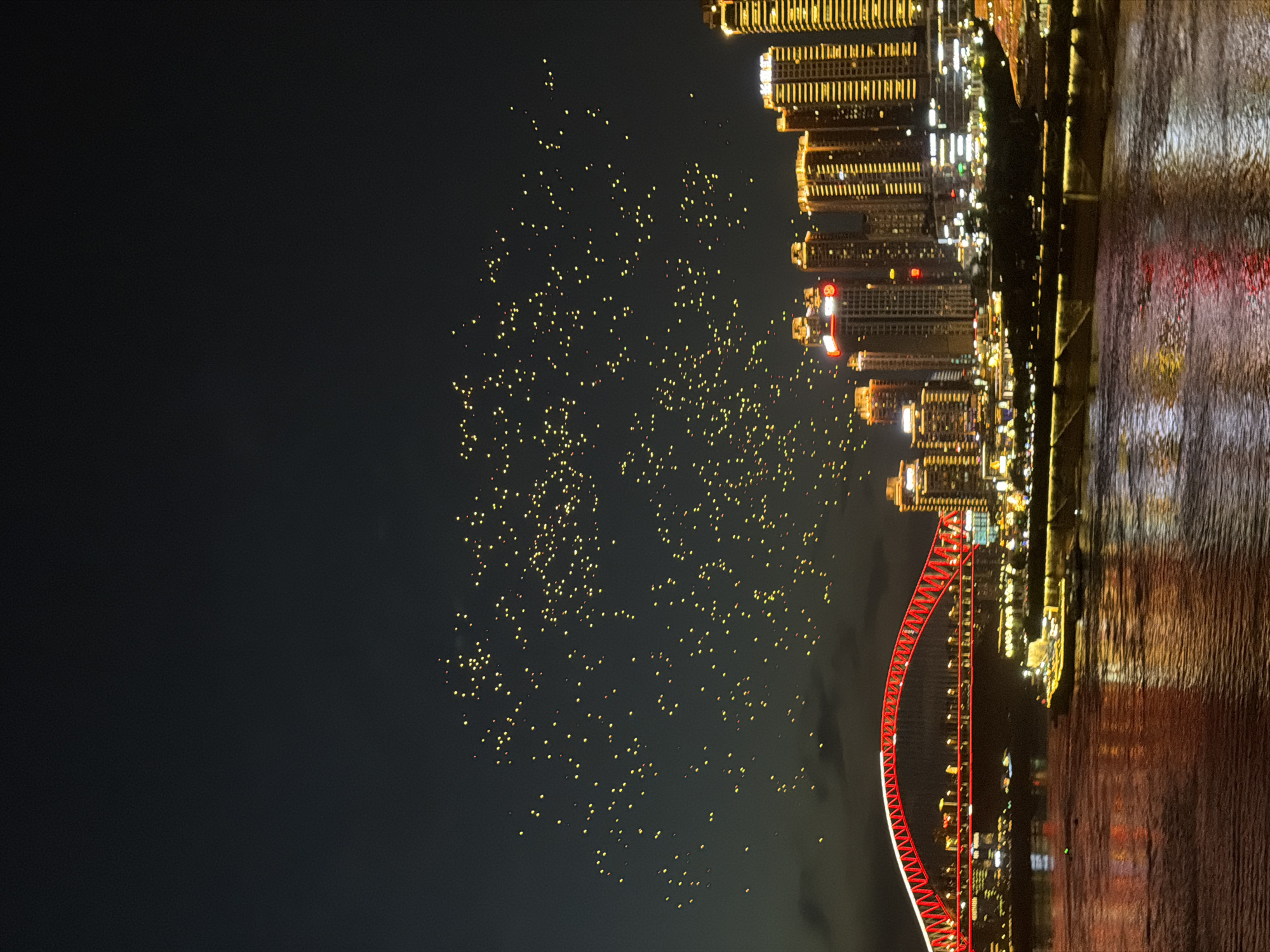 This is a nighttime photograph of a cityscape in Nan'an District, China, featuring a body of water in the foreground reflecting the city lights. A large, illuminated red bridge with white lighting along its arch spans across the water. Beyond the bridge, a dense cluster of tall buildings is visible, illuminated with warm yellow lights. In the sky above the city, numerous small, yellow lights are scattered, appearing to be sky lanterns or drones. The sky is dark, indicating it is nighttime, with some faint clouds visible. Several boats and structures are visible along the waterfront.