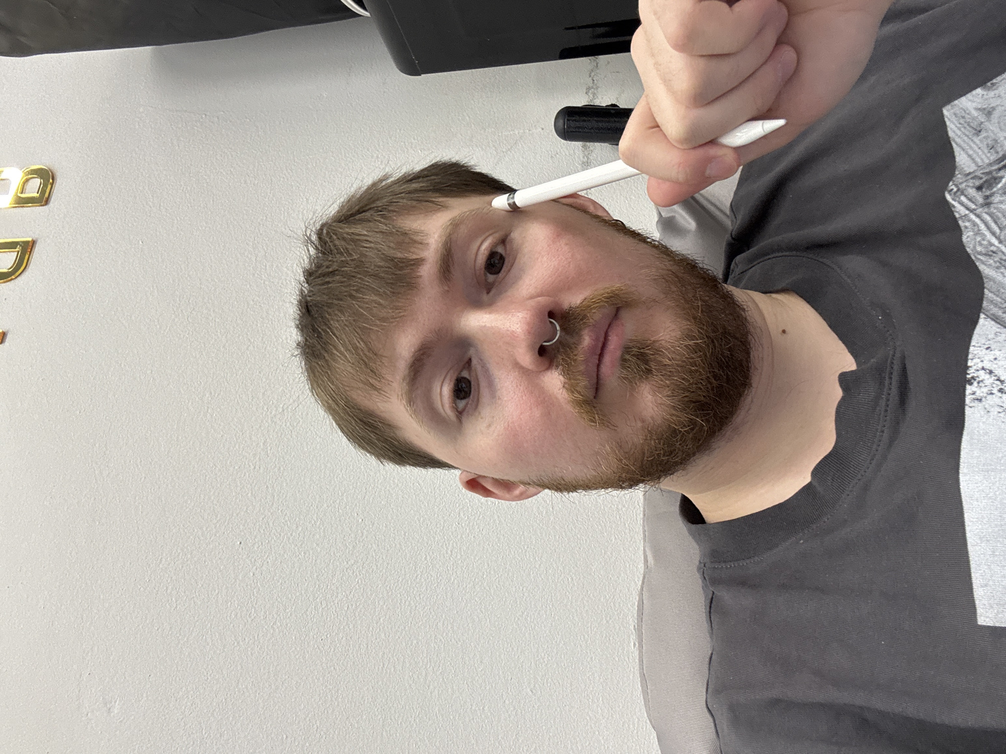 A man with light brown hair, a beard, and a nose ring holds a white stylus near his forehead. He is wearing a gray t-shirt. Behind him is a white wall with gold lettering and a black object. The location is unknown, with no information available to indicate the city of Chaikovsky, Russia.