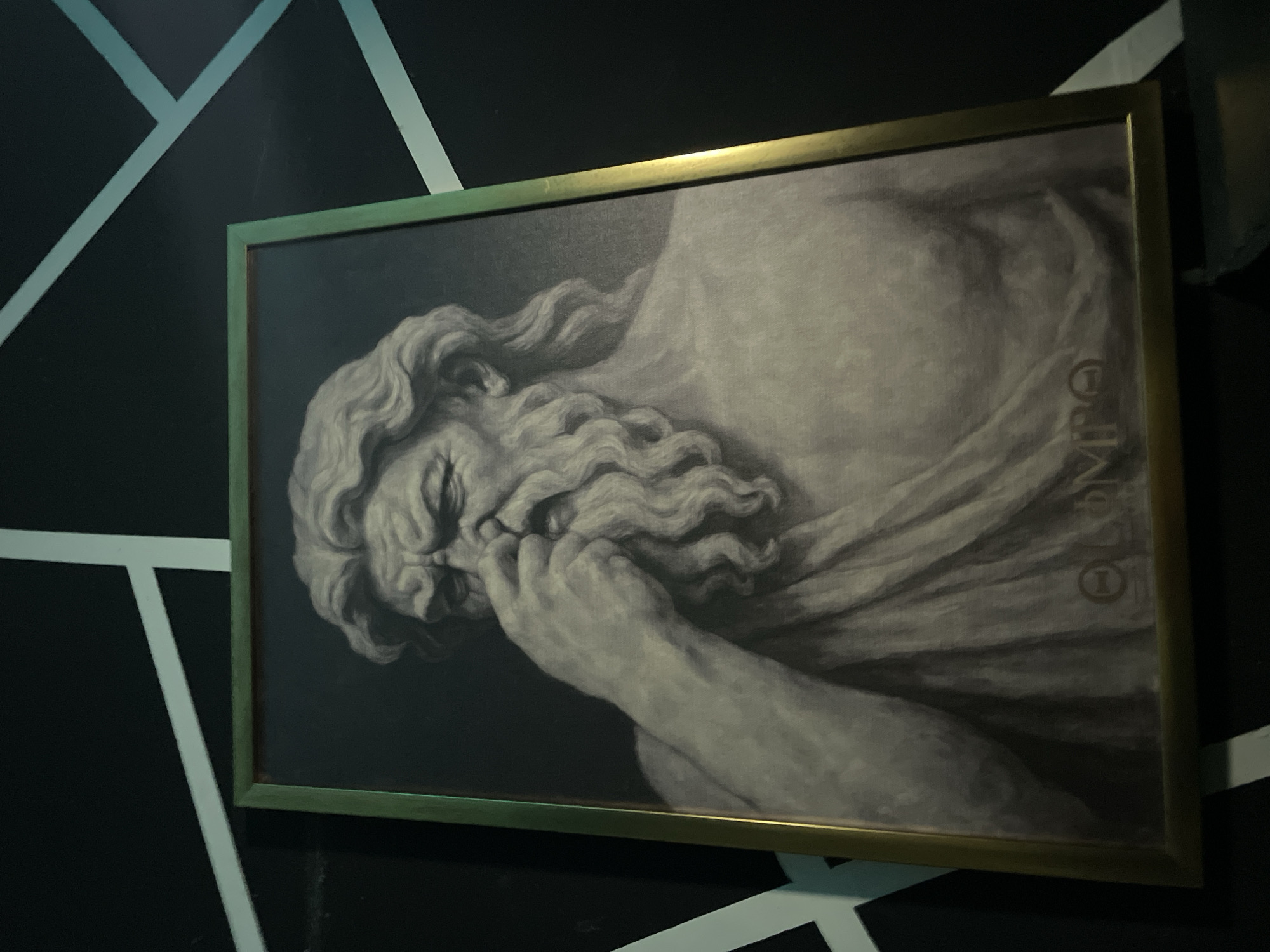 The image shows a framed black and white print of a classical-style portrait of an elderly man with long, flowing hair and beard, dressed in a draped garment. He is holding his hand near his face. Faint text, possibly "OLYMPUS", is visible on the right side of the print. The print is framed in a gold-colored frame and hangs on a wall decorated with a black and white geometric pattern of angled lines. The location is Mexico City, Mexico.