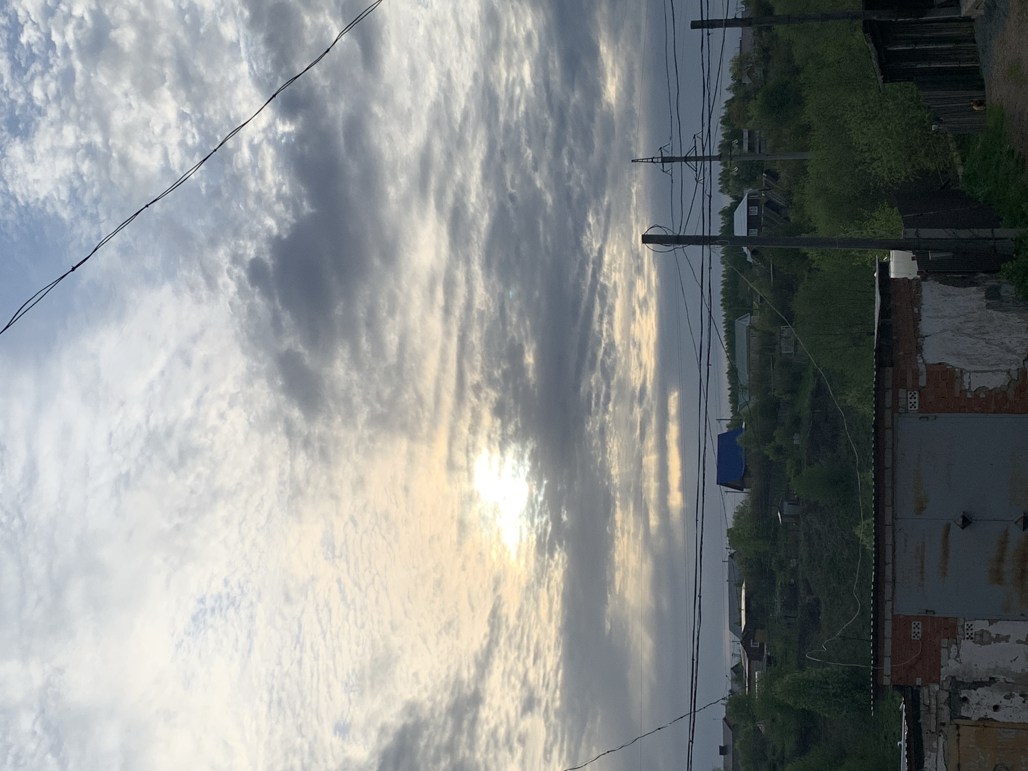 This image, taken in Фоки, Russia, captures a cloudy daytime sky above a semi-rural landscape. The sun is a dominant feature, shining brightly through a break in the clouds, casting a warm, luminous glow across the sky. The clouds exhibit various patterns, including streaky and rippled formations.
In the mid-ground and foreground, several utility poles, both wooden and concrete, crisscross the scene with numerous overhead electrical or communication wires. One pole on the left features an attached street lamp. The lower portion of the frame is filled with abundant green trees and bushes, suggesting a lush, possibly late spring or summer environment.
Among the vegetation, several buildings are visible. On the right, a weathered structure, likely a garage or an old shed, stands prominently with exposed brickwork and peeling white plaster or paint. Its roof appears to be made of corrugated metal. Further in the background, other residential buildings, including one with a distinct blue roof and another with a white facade, are nestled amongst the trees. A faint dirt path is visible towards the far right.
The scene depicts no specific activity, offering a static, naturalistic view of a village or settlement. The time of day is clearly daylight, indicated by the sun's bright presence. No text is visible in the image.