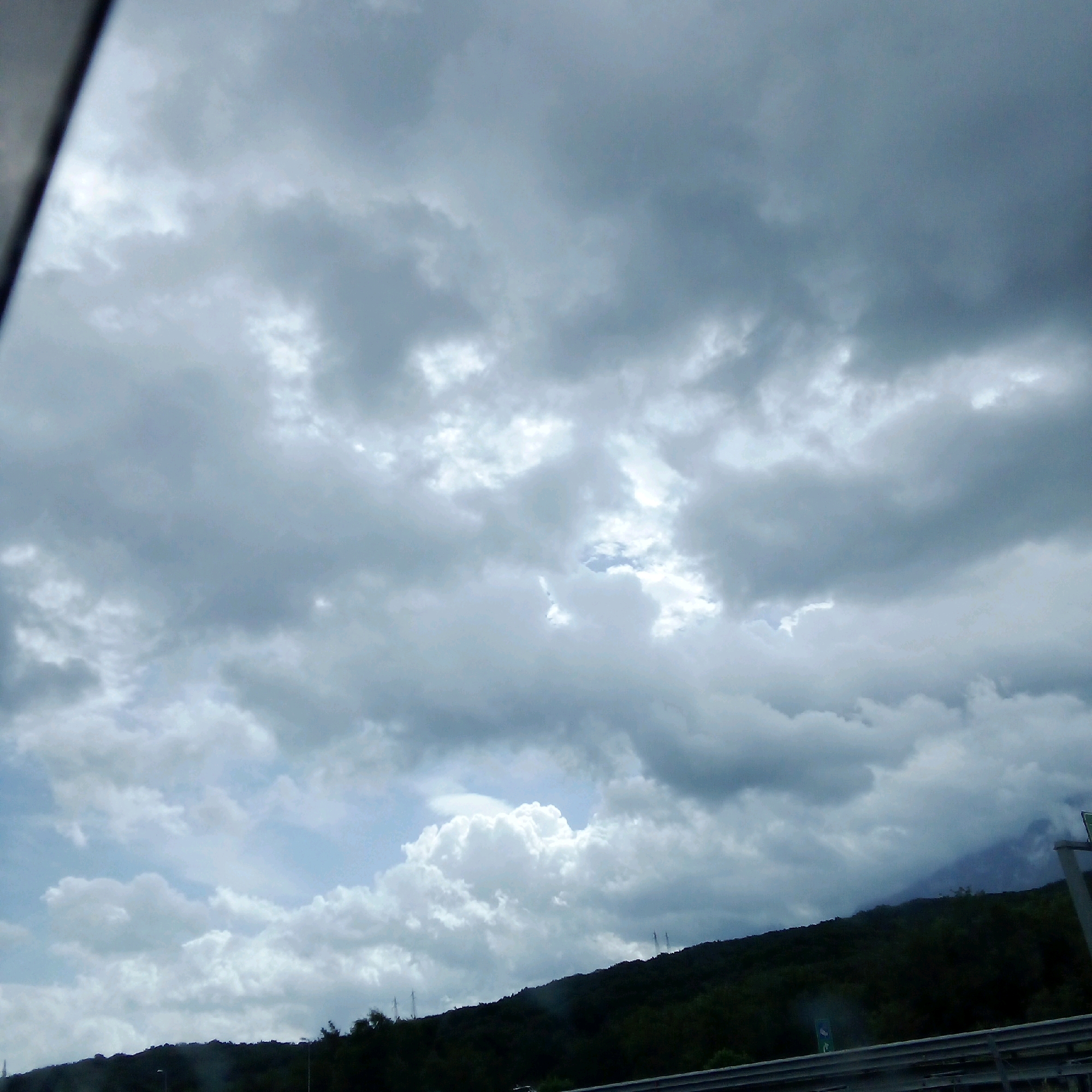 The image captures a dramatic sky filled with heavy, gray clouds. A break in the clouds in the upper center of the frame reveals a patch of bright blue sky. The lower portion of the image shows a dark, densely forested hillside, likely a mountain, silhouetted against the sky. Several utility poles or towers are visible on the hillside. In the foreground, a metal guardrail suggests a road or elevated viewpoint. The scene is overcast and potentially stormy. This view is consistent with the mountainous terrain surrounding Postiglione, Italy, a town located in the province of Salerno, Campania.