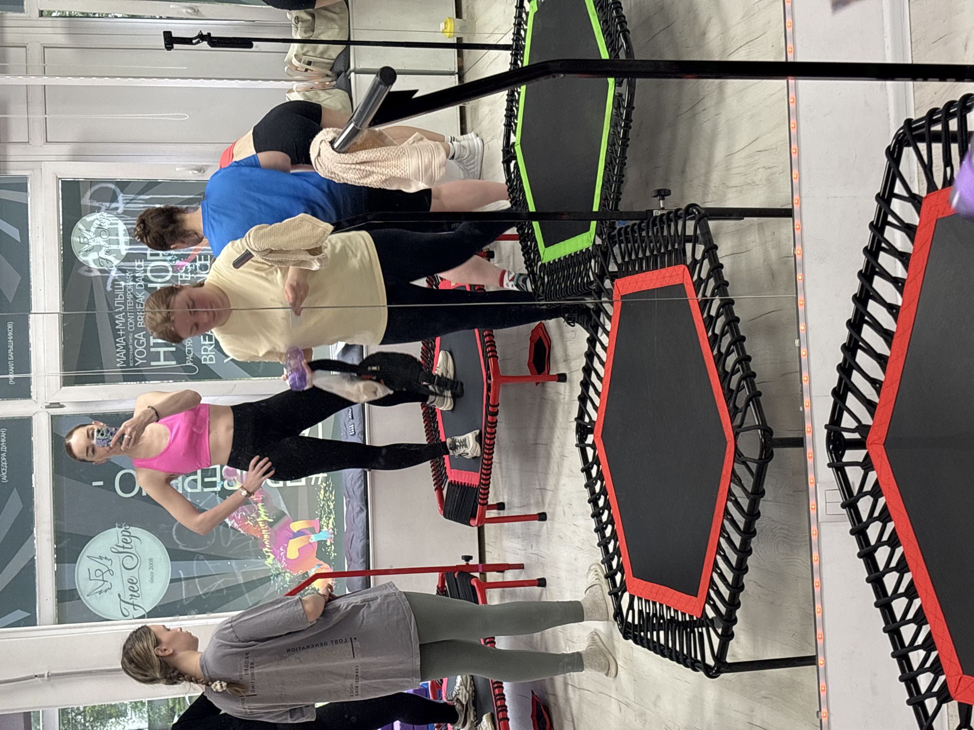The image appears to be taken indoors, possibly in a fitness studio or gym in an unidentified city in Russia, as suggested by the Cyrillic text in the background. Several individuals are present, engaged in fitness activities. One woman wears a pink top and black leggings and appears to be taking a selfie with her phone. Another woman, dressed in grey activewear, stands nearby. Other individuals are positioned near small trampolines with support bars, some of which have black surfaces with red or green edges. The trampolines are arranged in a line. The walls feature signage with text in both English and Russian, including the phrase "Free Steps since 2008" and other words related to yoga, break dance, and contemporary dance. The environment seems to be set up for a group fitness class.