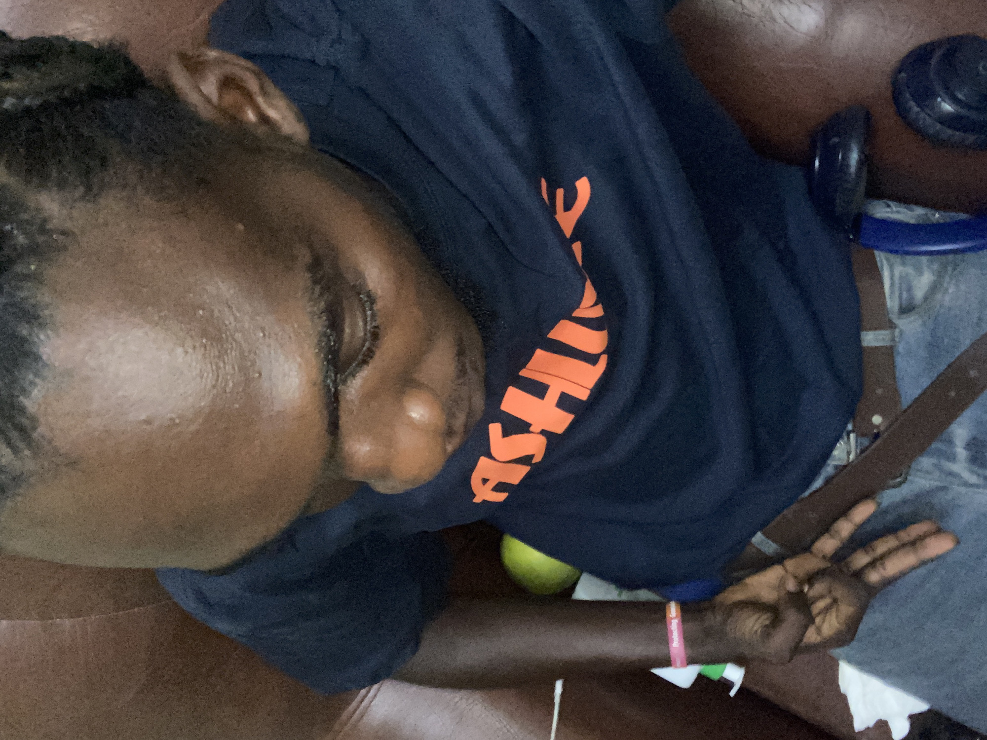 An overhead medium shot depicts a dark-skinned male individual seated on a brown, upholstered chair or sofa. The individual wears a dark blue t-shirt with partially visible orange text 'ASHI' on the chest, light blue denim jeans, and a brown leather belt. His hair is styled in short braids, and his forehead exhibits visible perspiration and skin texture. The subject's gaze is directed downwards and to the viewer's left. His left hand, adorned with a pink and white wristband (partial text 'Protecting Gen...'), rests on his lap, forming a V-sign with the extended index and middle fingers. A crumpled white tissue is visible on the jeans beneath the left hand. To the individual's left, a spherical yellow-green object, consistent with a fruit, is present. To the right, a dark blue object resembling a component of a snorkeling mask is partially visible. The scene is described as being located in Lagos, Nigeria, though no specific geographical identifiers are discernible within the image frame.