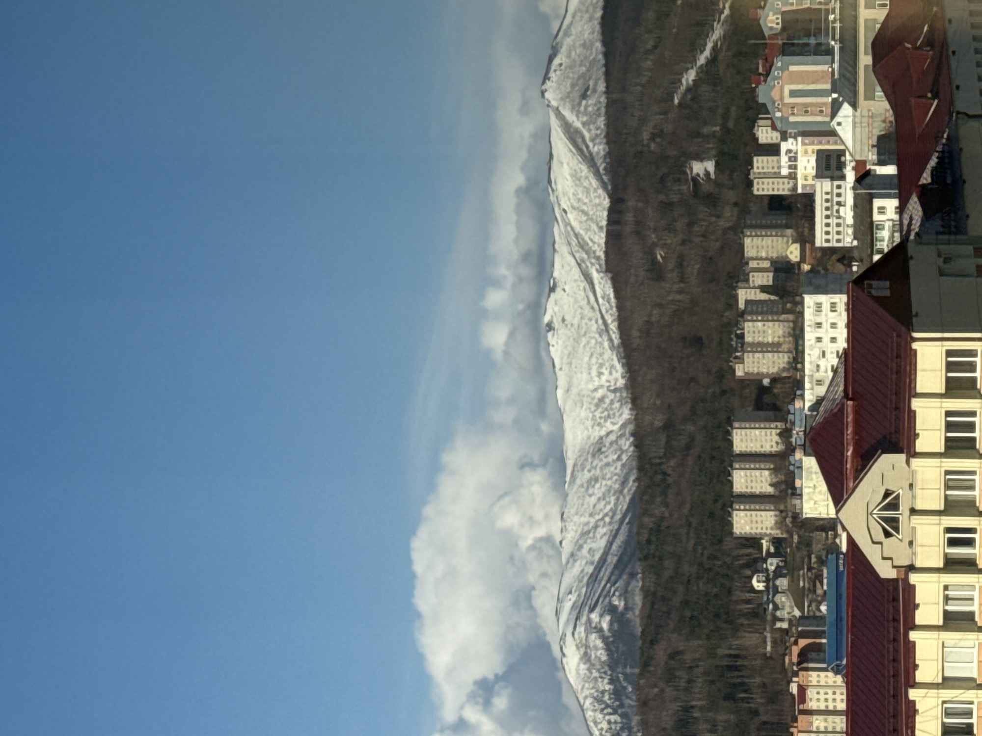 The image displays a panoramic view of an urban landscape situated against a backdrop of snow-capped mountains under a clear sky. In the upper portion, a vast expanse of bright blue sky is visible, dotted with scattered white clouds, including a more pronounced, elongated cloud formation running parallel to the mountain ridge.
The mid-ground is dominated by a majestic mountain range, its upper slopes and peaks covered in a thick blanket of white snow. Darker areas, likely exposed rock or evergreen trees, peek through the snow. Below the snowline, a dense, dark forest occupies the lower mountain slopes and extends towards the foreground, creating a natural boundary with the city.
The foreground and right side of the image feature a bustling urban settlement. Numerous multi-story buildings, predominantly in light beige or cream colors, populate the area. Many structures exhibit typical architectural elements such as rectangular windows and balconies. A striking feature is the prevalence of deep red, often steeply pitched roofs, which contrast sharply with the lighter building facades. One building in the immediate foreground showcases a decorative gable with a unique window design. The urban area appears to be a residential or mixed-use zone, with blocks of buildings extending into the distance towards the forested foothills.
The scene is set during a clear and bright day, likely late morning or early afternoon, indicated by the crisp lighting. The weather appears cold, as evidenced by the snow on the mountains. No people or specific activities are visible, emphasizing the serene yet developed environment. The combination of snowy mountains and a dense urban area is consistent with locations like Yuzhno-Sakhalinsk, Russia. No discernible text is present in the image.