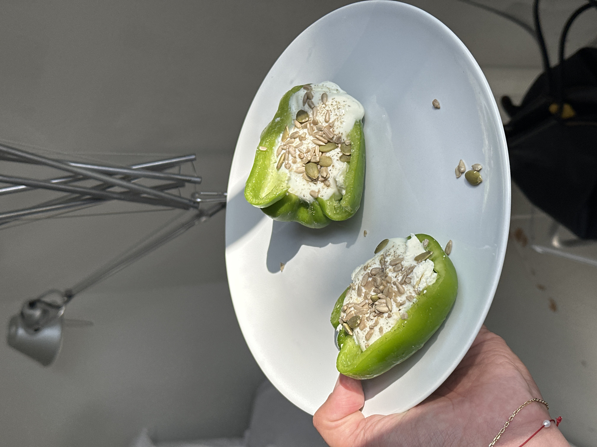 The image shows a hand holding a white plate with two halves of a green pepper stuffed with a white filling and topped with seeds. The plate also has a few loose seeds scattered on it. In the background, there is a chrome articulating lamp and a white surface. The location is Unknown, Unknown.