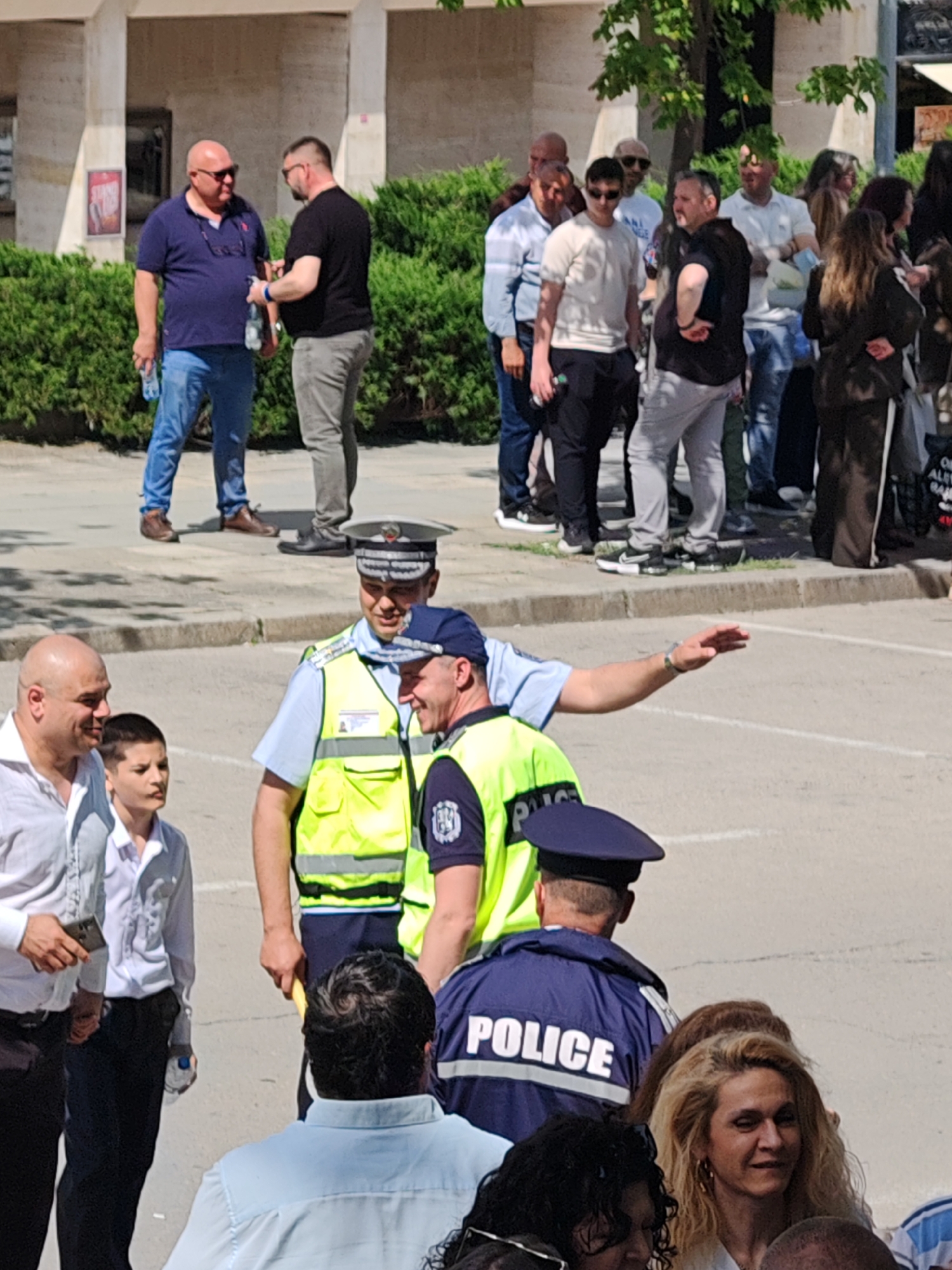 The image depicts a street scene in Shumen, Bulgaria. A small crowd of people is gathered near a building with white columns. Two men stand in the foreground; one is bald and wears a white shirt, and the other is a boy wearing a white collared shirt and black pants. Behind them are two police officers wearing light blue shirts and yellow reflective vests. One officer is gesturing with his right arm. Another police officer stands facing away from the camera, and his jacket reads "POLICE." The ground is paved with light gray stone. In the background, additional people are standing, including two men talking and holding water bottles. A row of green bushes is visible behind the men.