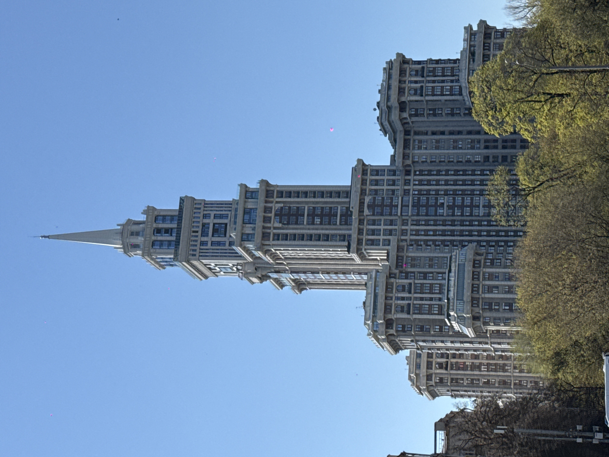 The image depicts a tall building with a spire against a clear blue sky. The building has a stepped design with multiple tiers. Its facade features a grid of windows. Trees are visible to the right of the building. The location is not in Khimki, Russia.