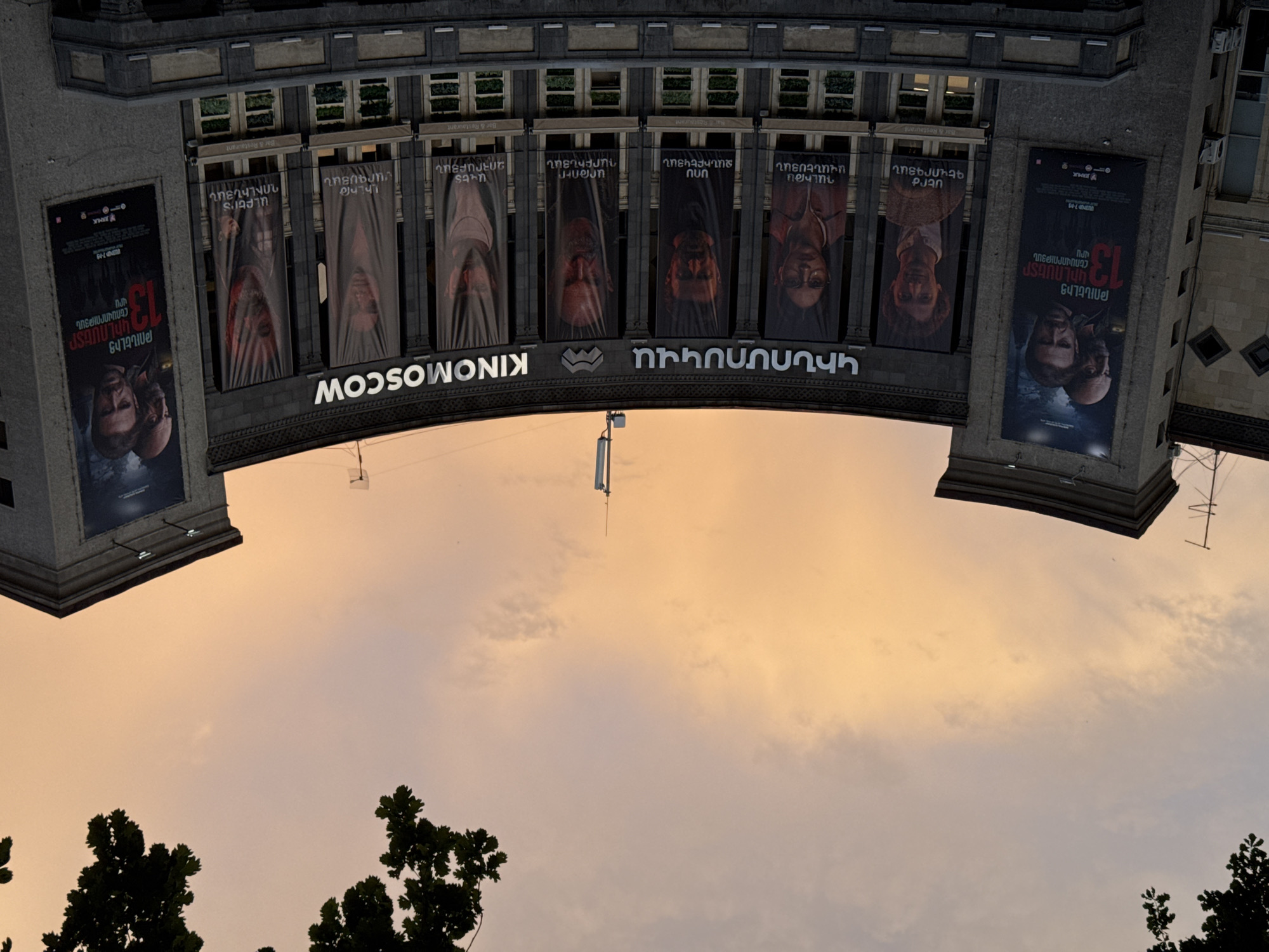 The image shows the Kinomoskva cinema in Yerevan, Armenia, viewed from a low angle, giving the impression that the cinema is inverted against the evening sky. The sky is a blend of pale orange and light gray, suggesting either sunrise or sunset. The building's facade is stone or concrete, with a row of windows and vertical banners displaying images of people. Two large movie posters are affixed to the corners of the building. The cinema's name is prominently displayed in Armenian. Trees frame the bottom of the shot. A thin white object, perhaps a flagpole or lighting fixture, is visible.
