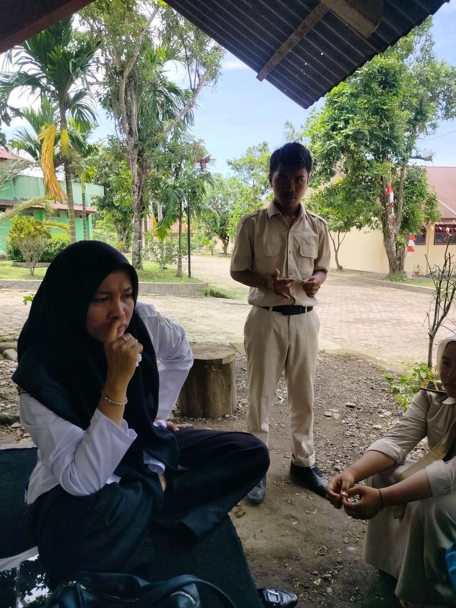 In Purwodadi, Indonesia, two young adults, a female wearing a black hijab and white uniform, and a male in a tan uniform, are outdoors. The female is seated, looking thoughtful with her hand near her face. The male stands behind her, holding a small object between his fingers, appearing to gesture or explain something. A third person, partially visible and also in uniform, is seated to the right. They are in an open area with trees, foliage, and a building in the background, suggesting a school or public space. The ground is covered with gravel and dirt, with a tree stump nearby. A corrugated roof structure is visible overhead.