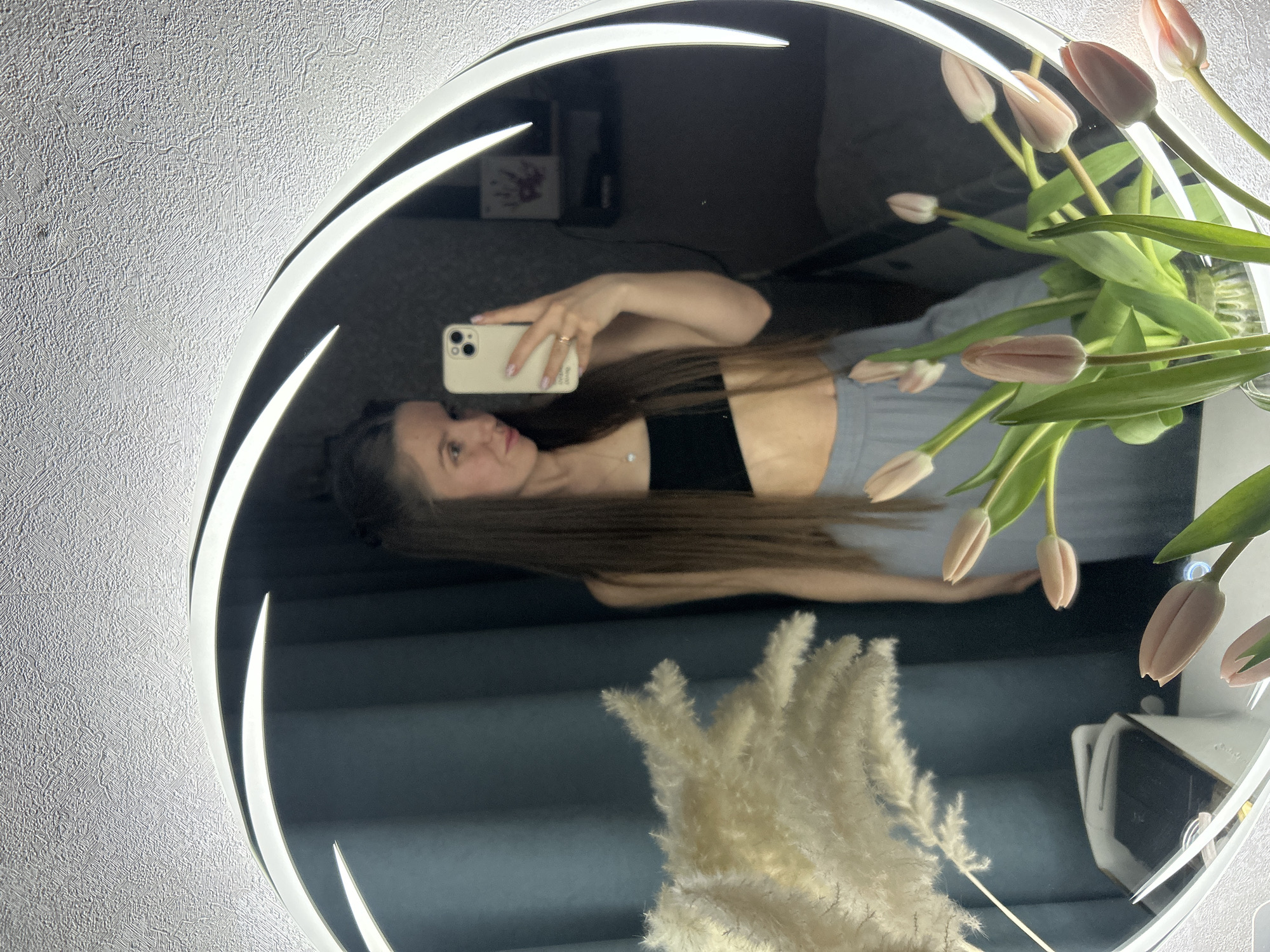 The image depicts a reflection in a circular mirror with an integrated light. The background appears to be a textured wall. A young woman is visible in the mirror's reflection, holding a smartphone and taking a selfie. She is wearing a black top and grey bottoms. Part of a floral arrangement, including pink tulips and dried pampas grass, is reflected in the mirror. In the background, a picture frame is faintly visible. The city of Klin, Russia, is not identifiable as the location from the given image.