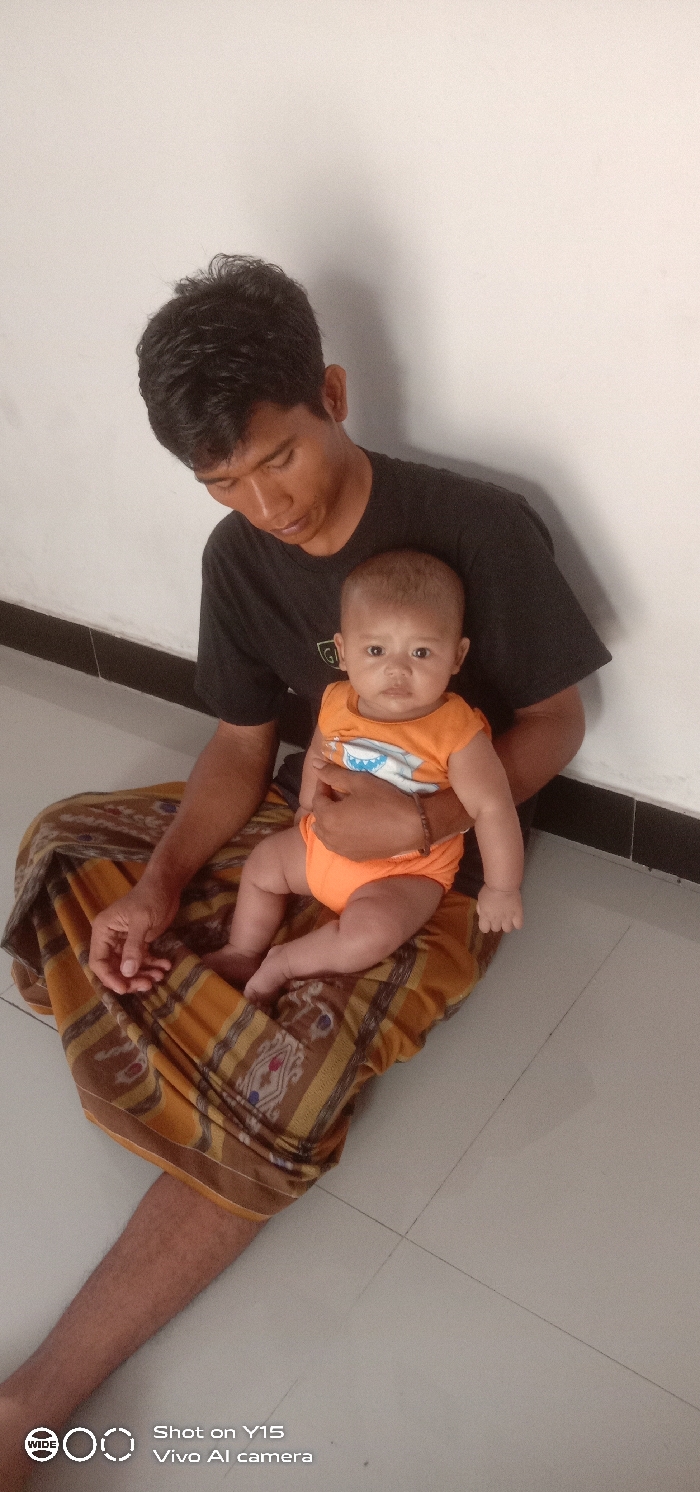 An adult male is seated on a light grey tiled floor against a plain white wall with a black skirting board, holding an infant in the city of Brumbungan Kidul, Indonesia. The adult male has short dark hair, a dark complexion, and is wearing a black short-sleeved t-shirt and a patterned sarong in shades of brown, orange, and gold. His gaze is directed downwards. He supports the infant with his left arm while his right hand rests on his upper leg. The infant, with light brown skin, wears a sleeveless orange onesie featuring a white graphic on the chest. The infant is positioned upright, facing directly towards the camera, with a neutral facial expression. The infant's legs are bent, and feet are bare. The scene is illuminated by ambient indoor light. A "Shot on Y15 Vivo AI camera" watermark is present in the lower left corner.