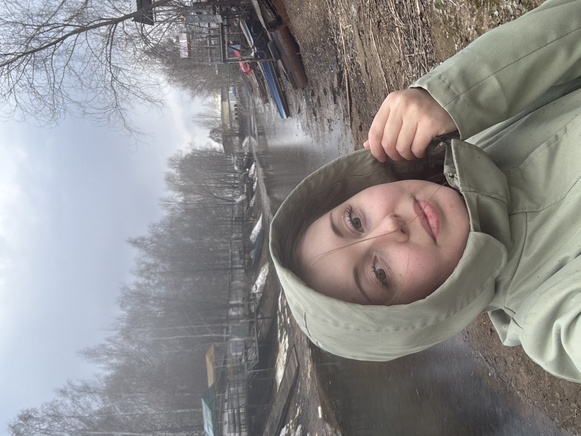 A young woman wearing a light green hooded jacket is facing the camera. The background features a river, trees, and what appears to be a waterfront area with buildings and boats. The location is Unknown, Unknown. The weather appears foggy or overcast.