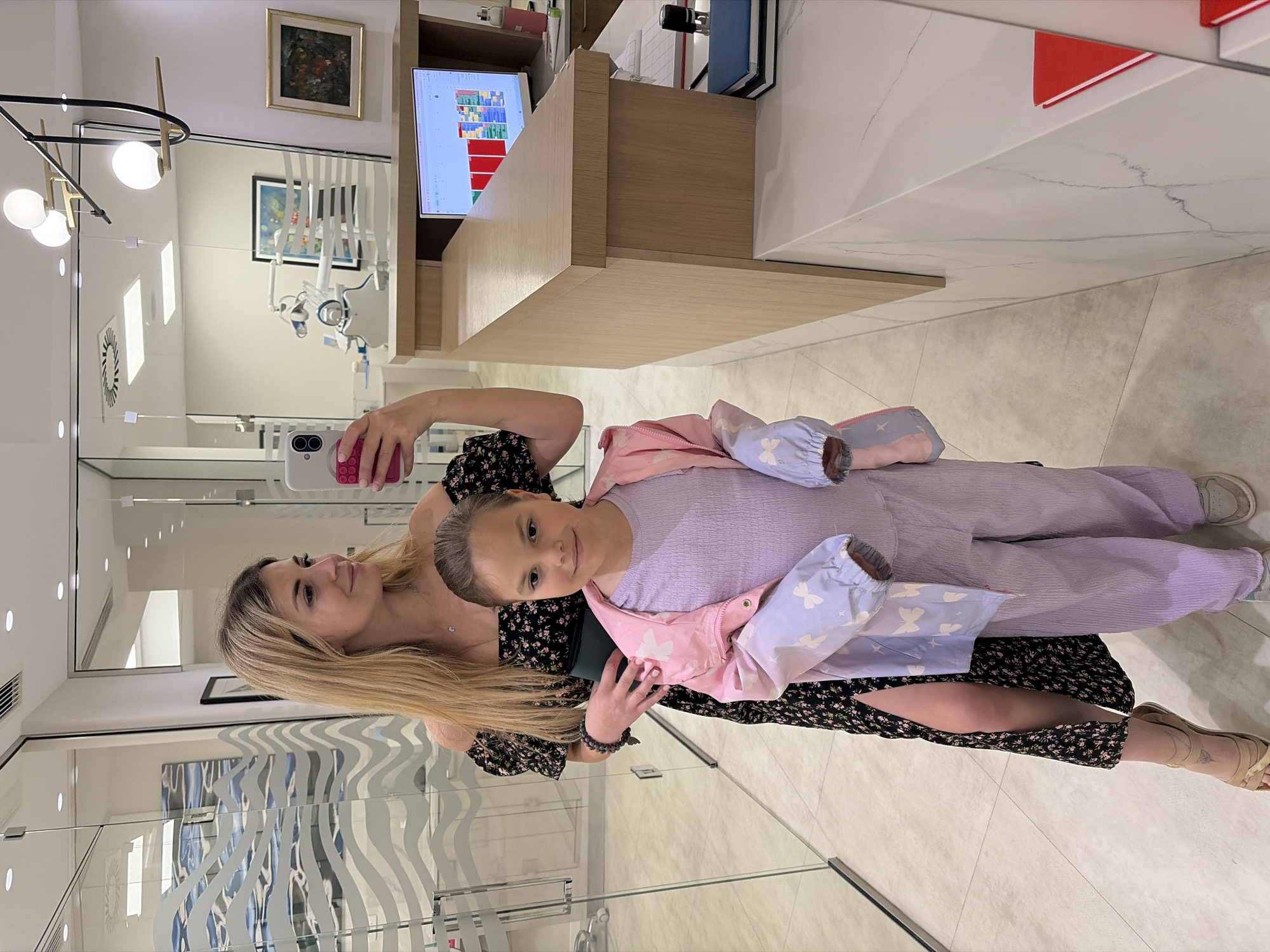 A fair-skinned woman with long blonde hair takes a photo with a pink cellphone of a fair-skinned child with her blonde hair pulled back into a bun in what appears to be a modern dentist's office in Budva, Montenegro. The woman wears a black dress with pink floral detailing. The child wears a lavender-colored jumpsuit with a light pink jacket with light blue sleeves adorned with white butterfly designs. The dentist's office has a flat-screen TV on the wall, dental equipment visible through a glass partition, modern lighting fixtures, and abstract artwork on the glass walls. The floor is tiled with a light-colored tile.