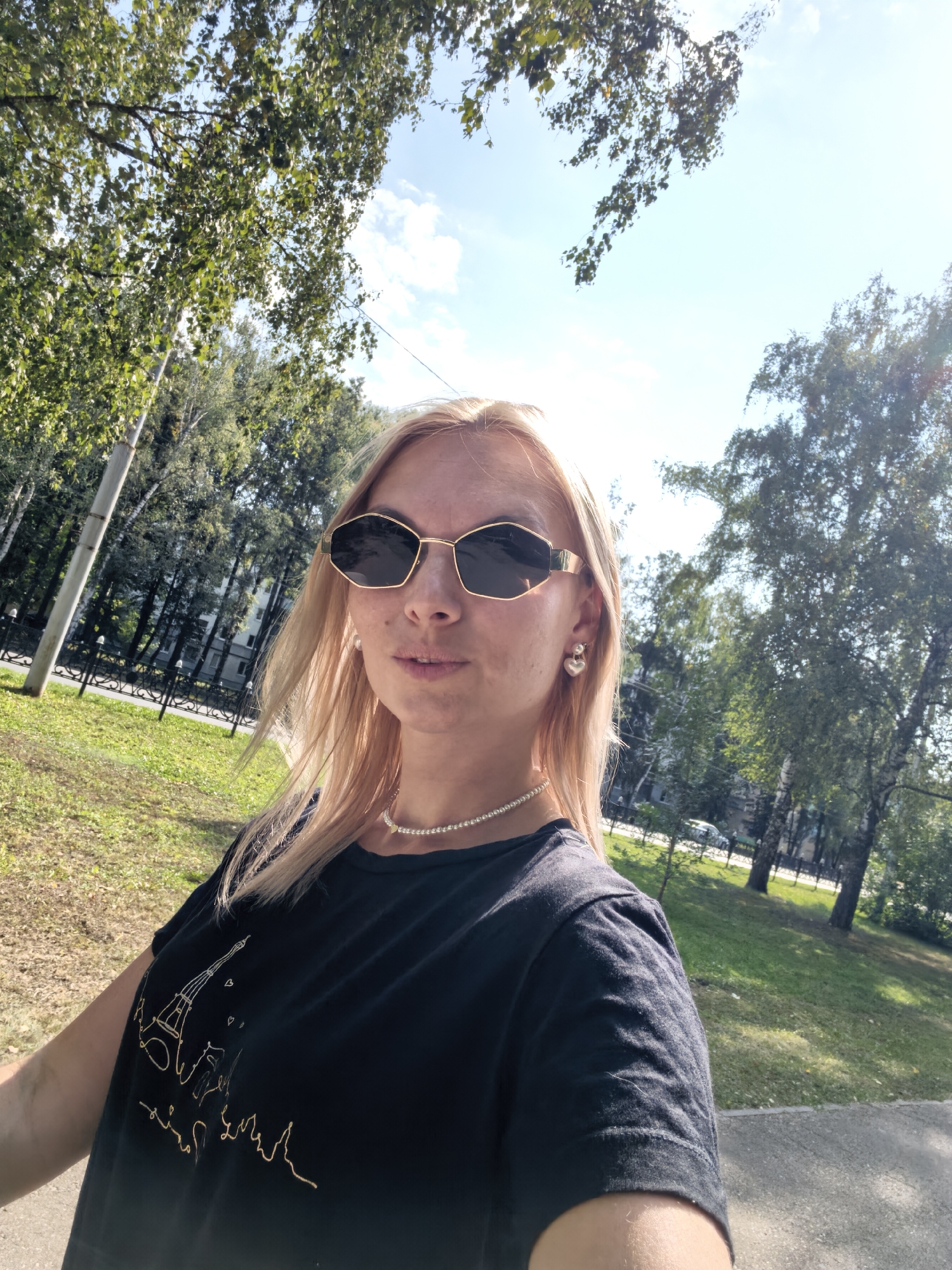 A woman with blonde hair and sunglasses takes a selfie outdoors. She is wearing a black t-shirt with a gold Eiffel Tower graphic, a pearl necklace, and heart-shaped earrings. The background shows trees, grass, a sidewalk, and a building in the distance. This image was taken in Sterlitamak, Russia.