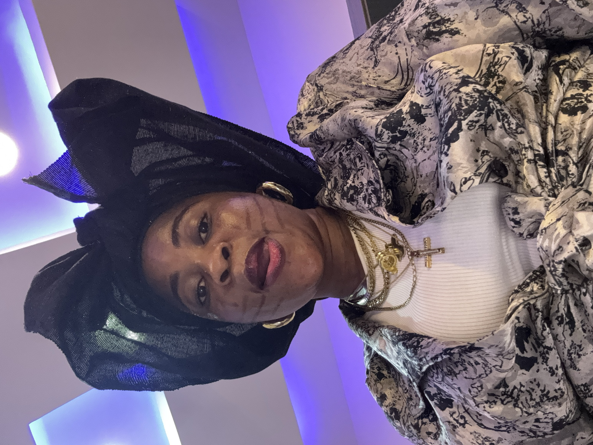 A medium-shot, front-facing photograph captures a Black woman indoors, likely in a residential setting in Ajara, Nigeria. She wears a large, black, textured headscarf and a white top layered with a grey and black patterned jacket. Multiple gold necklaces adorn her neck, including a cross pendant and a round pendant with a figure. She has large hoop earrings in her ears. The background features a ceiling with purple accent lighting. The woman is looking directly at the camera, with her mouth slightly open and her tongue visible. Her facial makeup includes dark lines drawn horizontally across her cheeks and forehead.