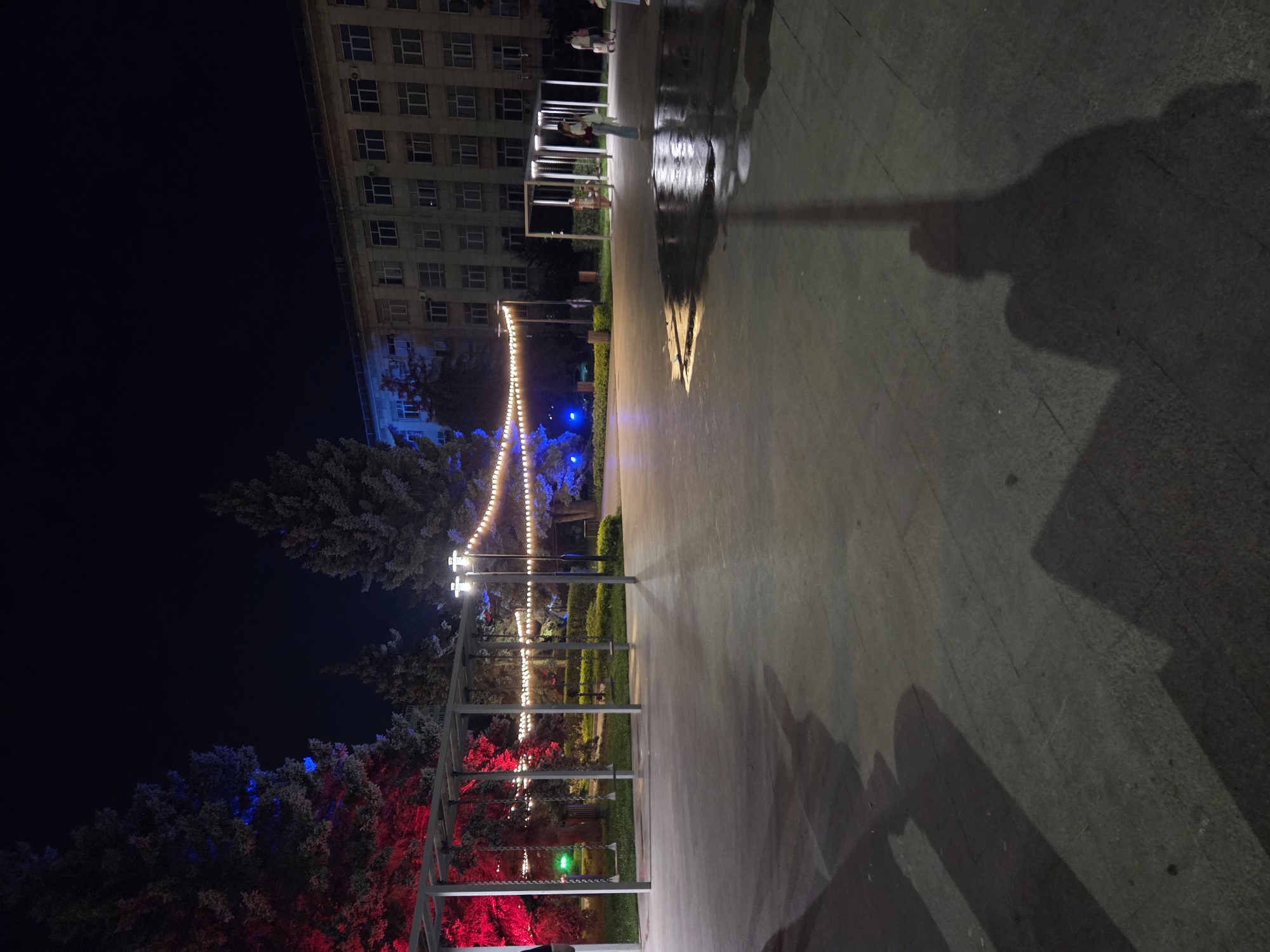 The image captures an outdoor public space at night in Chelyabinsk, Russia. The foreground consists of a paved area, possibly a plaza or walkway, with distinct shadows cast by unseen objects and people. In the middle ground, a park-like setting is visible, featuring trees illuminated with red and blue lights, string lights, and a structure with swings. A building, likely a government or educational institution given its architectural style with many windows, stands in the background, also illuminated. Several individuals are present in the middle ground, some near the swings and others walking. The overall impression is of a well-lit, active public area at night, potentially decorated for an event or holiday.