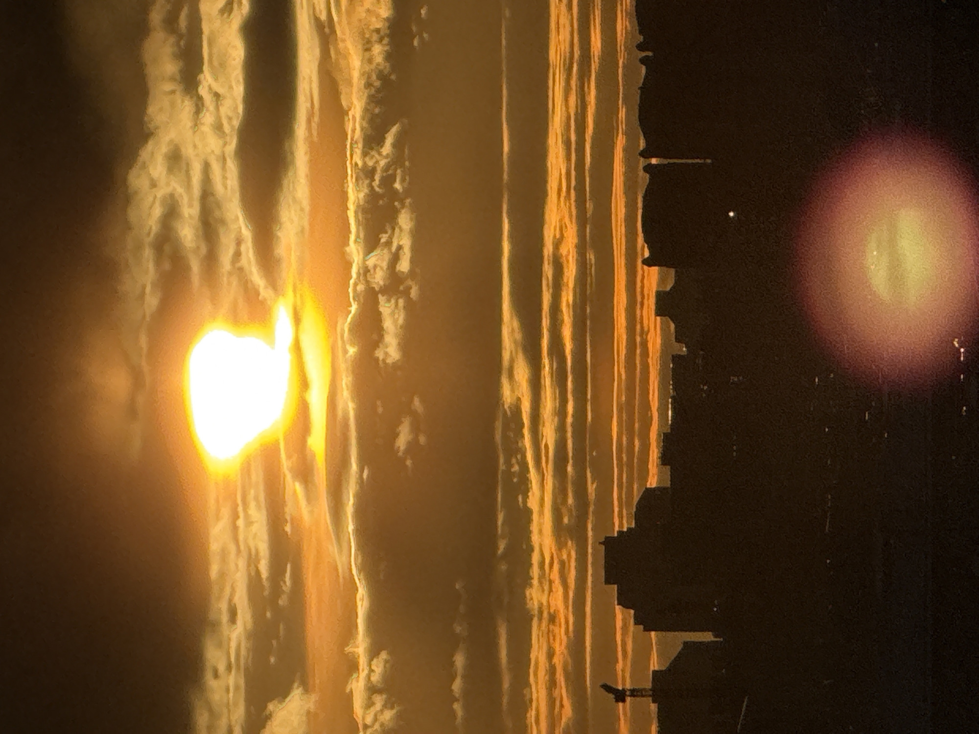 The image shows a sunset over what appears to be the skyline of Moscow, Russia. The sun, a bright yellow-white orb, is positioned on the left side of the image amidst clouds with a golden hue. The silhouette of buildings is visible against the darkening sky on the right. There is lens flare in the upper right of the image.