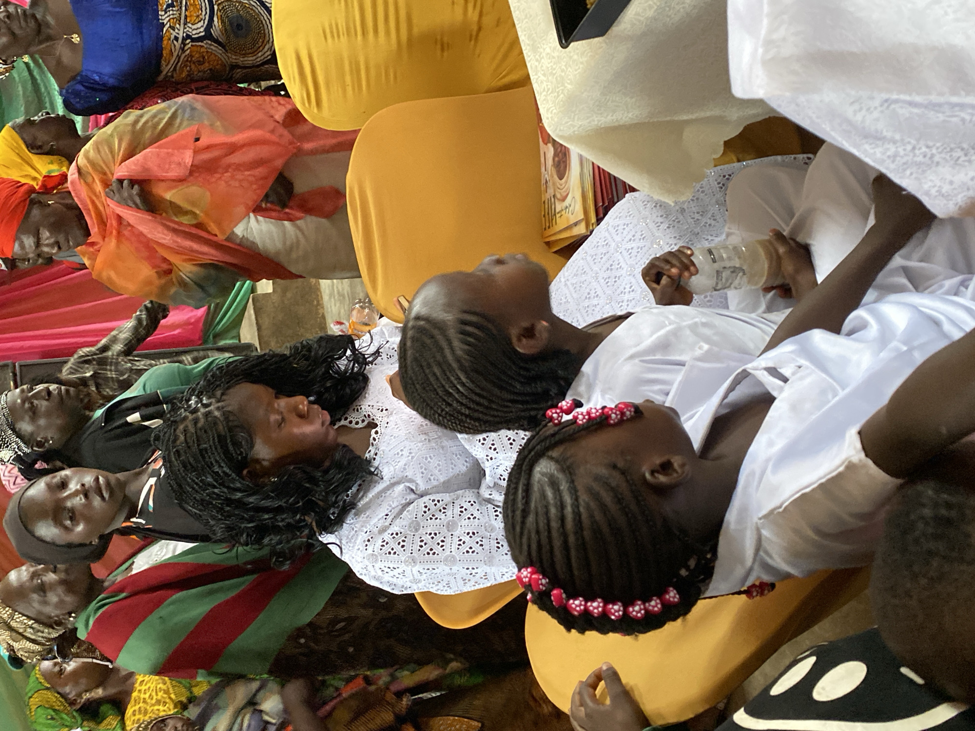 The image depicts a gathering of people, predominantly women and children, seated and standing indoors. The environment appears to be a community event or ceremony, with colorful fabrics draped in the background, suggesting a festive or formal occasion. Several individuals are wearing traditional African attire, characterized by vibrant colors and intricate patterns.
In the foreground, two young girls are prominently featured, both with braided hair. One girl wears a white lace garment and holds a bottle of liquid. The other girl, also in white attire, has her hair adorned with red and white beads. Behind them, an adult woman in a white lace top is visible. Further back, other women are seen, some wearing headscarves and draped cloths in shades of red, orange, green, and yellow.
The people are seated on yellow chairs and appear to be engaged in conversation or observing an event. The lighting suggests an indoor setting, possibly illuminated by natural and artificial light sources. The overall atmosphere is one of community and cultural engagement. The presence of various fabrics and attire indicates a possible celebration or religious gathering. The image captures a moment of shared experience within this community.