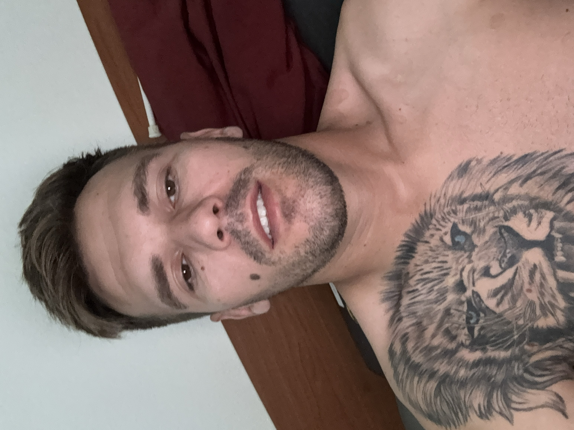 The image shows a close-up, rotated view of a man with a tattoo of a lion on his chest. He has short, dark hair, a beard, and a mole on his left cheek. His eyes are open and he appears to be smiling slightly, with his teeth visible. The man is shirtless, and part of his shoulder and chest are visible. In the background, there is a dark red fabric, possibly clothing or bedding, and a wooden surface that could be furniture. The lighting suggests it is daytime. The man is likely indoors, and the overall impression is casual and relaxed. The location context is Sesto Calende, Italy.
