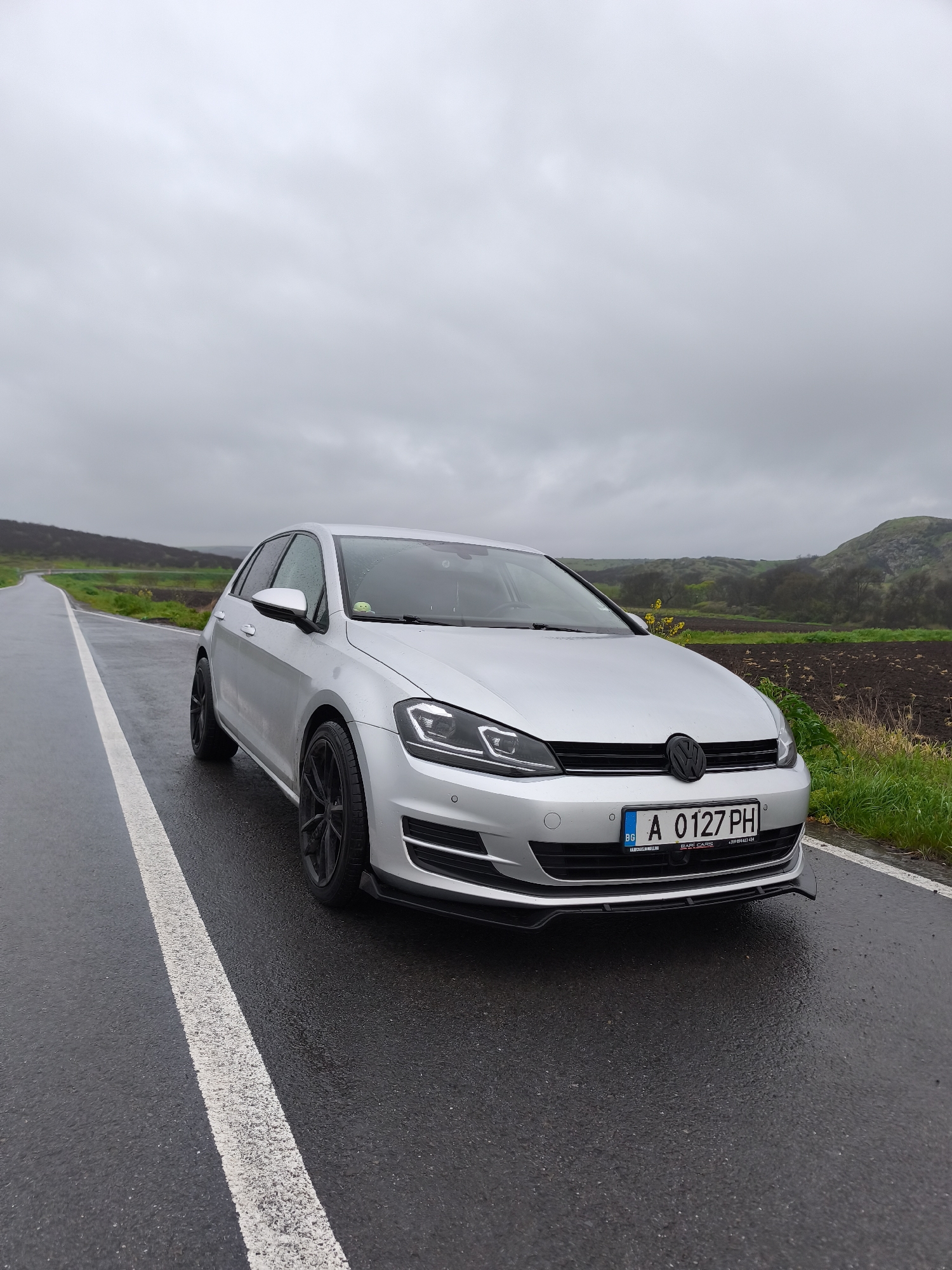 A silver Volkswagen Golf hatchback is parked on the side of a wet, asphalt road under an overcast sky. The car is angled, showcasing its front and left side, and features black multi-spoke alloy wheels and a black front bumper lip/splitter. A Bulgarian license plate, "A 0127 PH," is prominently displayed on the front, with "BG" indicating the country. Below the license plate, a black frame includes the text "BARI CARS" and a phone number (+359 894 633 434), along with a partially obscured word, possibly "KANALA NIMELHO," on the left. A small green plush toy is visible on the car's dashboard through the windshield.
The road is marked by a solid white line on its edge, leading into the distance. The wet surface indicates recent rain or damp conditions. The background reveals a rural or semi-rural landscape consistent with Rusokastro, Bulgaria, featuring gently rolling green hills and areas of dark, freshly tilled earth. Small yellow flowers can be seen among the roadside vegetation. The heavy, uniform grey cloud cover suggests a cloudy day with no direct sunlight, contributing to a muted and calm atmosphere. No people or active events are depicted, making the car the sole focus of the scene amidst the tranquil natural setting.