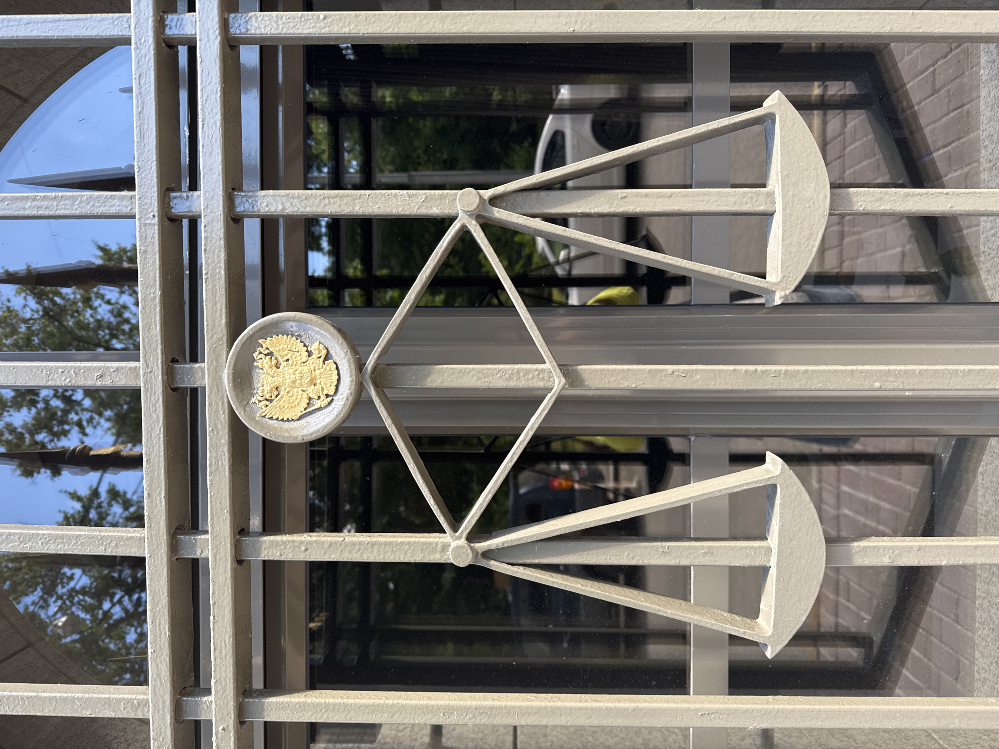The image shows a close-up of a metal gate in Moscow, Russia. The gate is painted a light beige color and features decorative elements. Prominently displayed is a gold emblem of the Russian double-headed eagle within a circular frame, positioned centrally above an artistic rendering of a balance scale. The scale is also made of beige metal and is designed symmetrically, with two suspended pans. The gate is set against a backdrop of a modern building entrance, characterized by large, reflective glass doors and windows. Through the glass, one can see trees, the sky, and the reflections of vehicles, suggesting an urban environment. The overall impression is one of officiality and possibly a governmental or judicial building, given the symbolic imagery of the eagle and the scales of justice.
