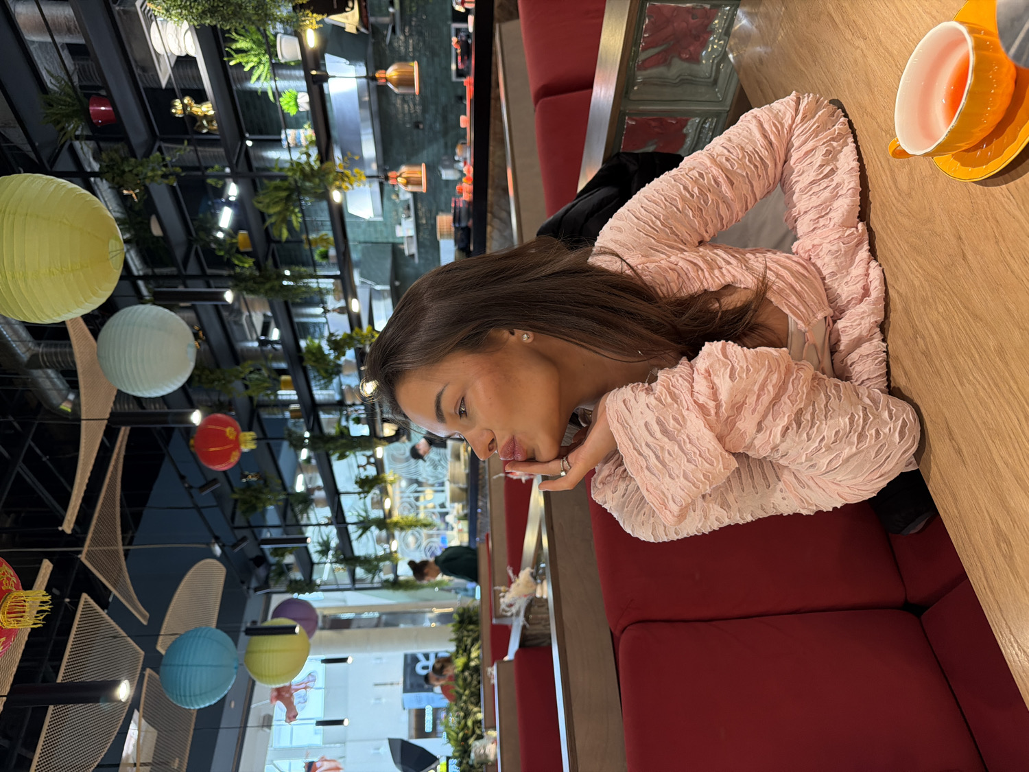 A young woman is depicted in an indoor restaurant or cafe setting in Krasnoyarsk, Russia. She is positioned at a wooden table with red booth seating, leaning on her forearms with her chin resting on her clasped hands. Her long, dark brown hair cascades over her shoulders, and she wears a light pink, textured long-sleeved top. Her expression is calm and contemplative, as she looks slightly away from the camera. An orange cup and saucer are visible on the table to her right.
The background reveals a modern interior with an industrial-chic aesthetic. Numerous colorful paper lanterns, in shades of yellow, light blue, red, and purple, hang from the dark, exposed ceiling structure. Lush green hanging plants are also interspersed throughout the ceiling, adding a natural element to the decor. Bright lights illuminate the space, suggesting a well-lit environment. Blurred figures of other patrons and elements of a service area are visible in the background, indicating a lively establishment. The overall scene conveys a relaxed dining or waiting moment. There is no visible, decipherable text in the image.