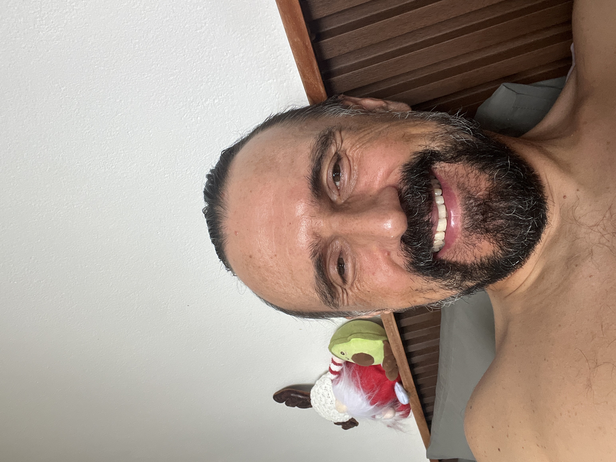 A man with a receding hairline, a black and gray beard, and visible wrinkles is smiling at the camera. He is partially shirtless, with his right shoulder visible. Behind him are a white wall, a wooden-slatted headboard, and a gray pillow. A decorative gnome with an avocado head, a white beard, and a red striped body is attached to the wall. The image appears to be taken indoors. There is no indication of the location being in บ้านบางรักษ์, Thailand.