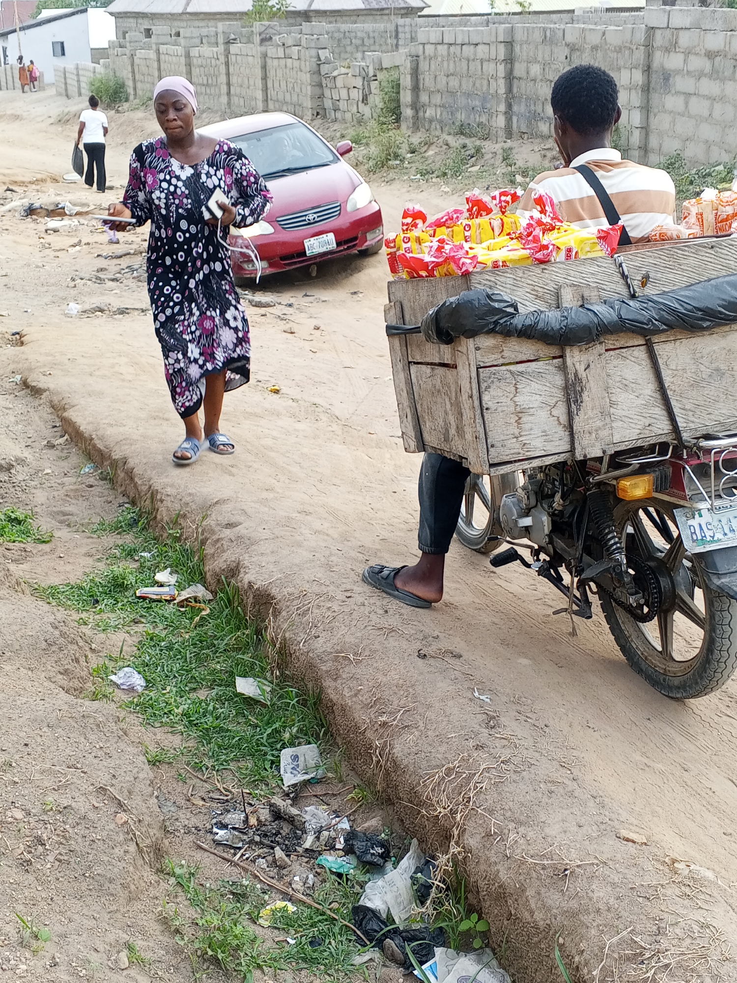 The image shows a street scene in Adonkolo, Nigeria. A woman wearing a floral dress and headscarf stands on a dirt path, holding what appears to be a phone and other small objects. Behind her, a red Toyota Corolla is partially visible. To her right, a man on a motorcycle is transporting a wooden box filled with packaged goods. The ground is dirt, with a ditch containing trash and vegetation along the side. In the background, there are buildings and other people. The motorcycle has license plate BAS-147-UA.
