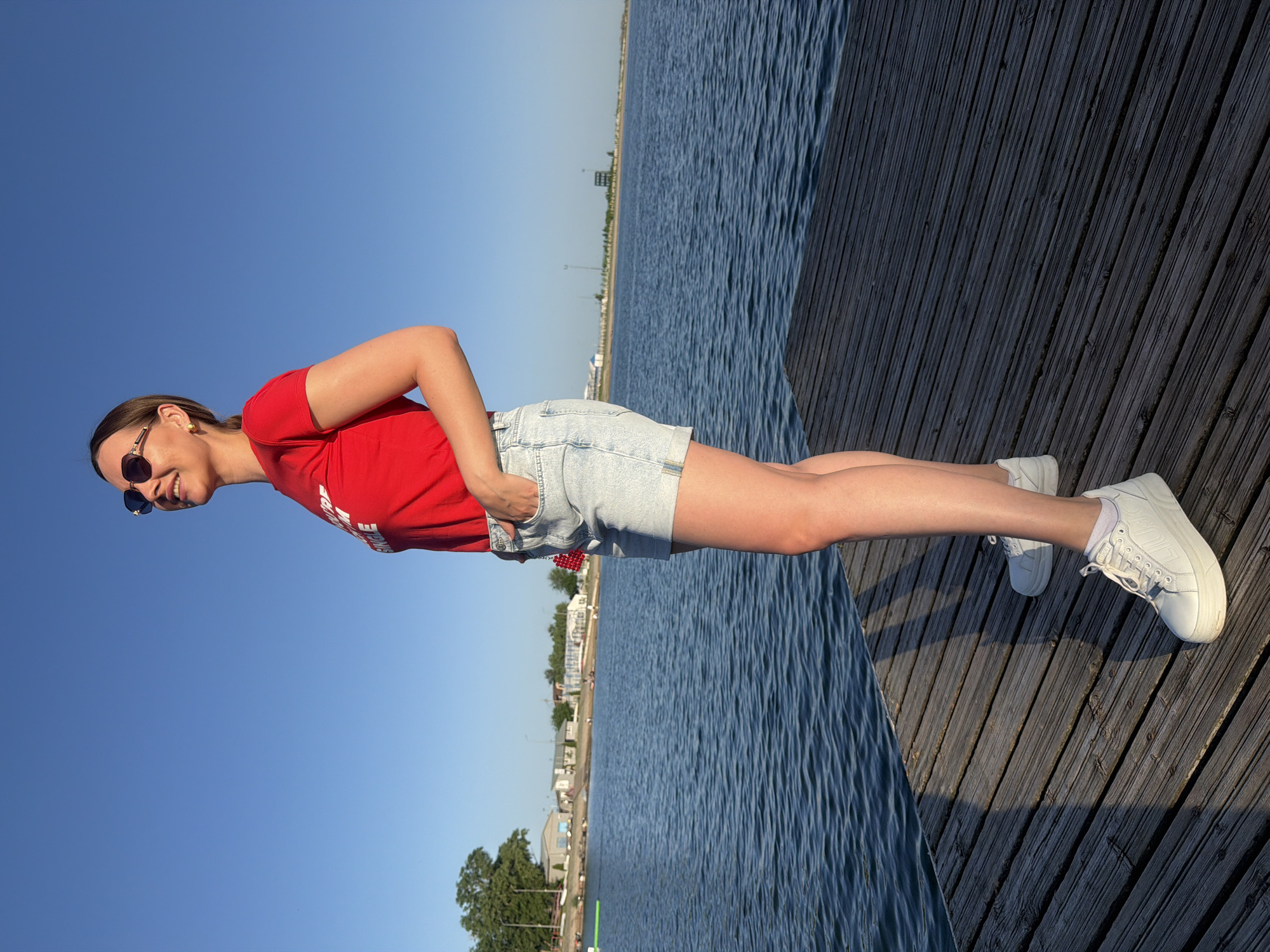 A woman stands on a wooden dock adjacent to a body of water, likely the Don River, in Rostov-on-Don, Russia. She is facing left, with her right hand in the pocket of her light blue denim shorts. She wears a red t-shirt and white sneakers. The sky is blue, and the background reveals a distant cityscape. The image is taken from a low angle and rotated 90 degrees counter-clockwise.