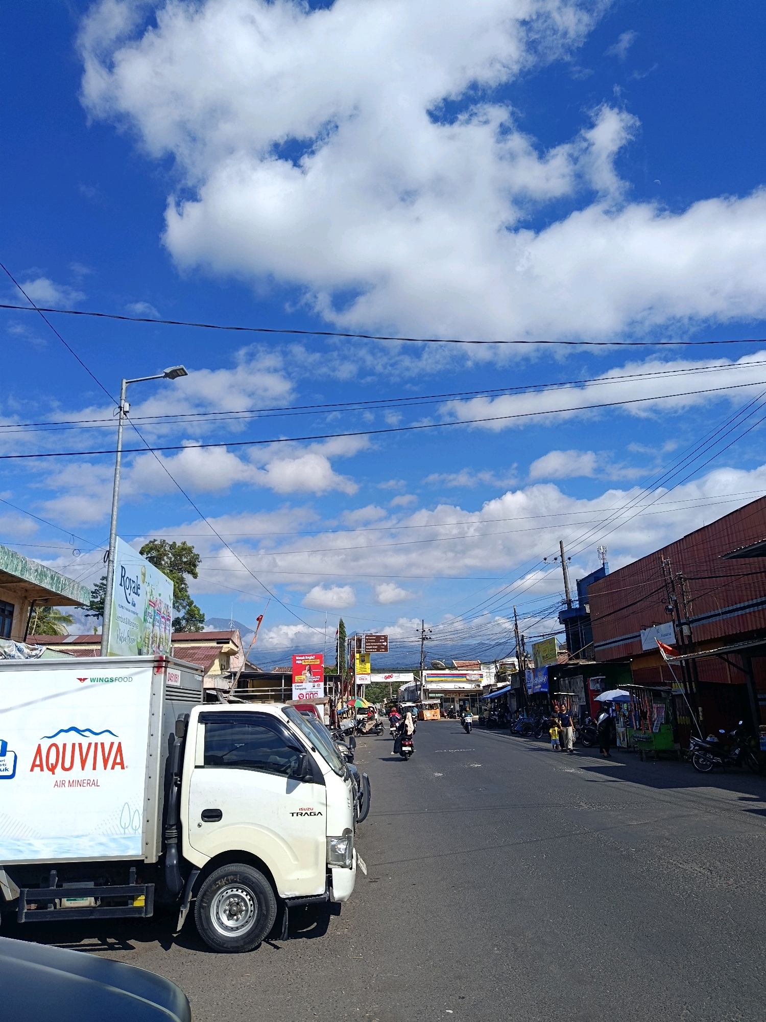 The image depicts a street scene in Senduro, Indonesia, on a bright, sunny day with a clear blue sky filled with scattered white clouds. In the foreground, the left side of the frame is dominated by the side of a white truck with "AQUVIVA AIR MINERAL" printed on its container. The truck is an Isuzu Traga. The road itself is paved and appears to be a main thoroughfare, with a line of shops and buildings lining the right side of the street. Several motorcycles are parked along the sidewalk in front of these establishments, and a few people are visible, including a woman and a child on the right side of the road, and other figures on motorcycles further down the street. A street lamp stands on the left side of the road, and numerous electrical wires are strung across the sky. In the background, mountains are faintly visible on the horizon, suggesting a rural or semi-rural setting within Senduro. The overall impression is of a typical day in a small Indonesian town.