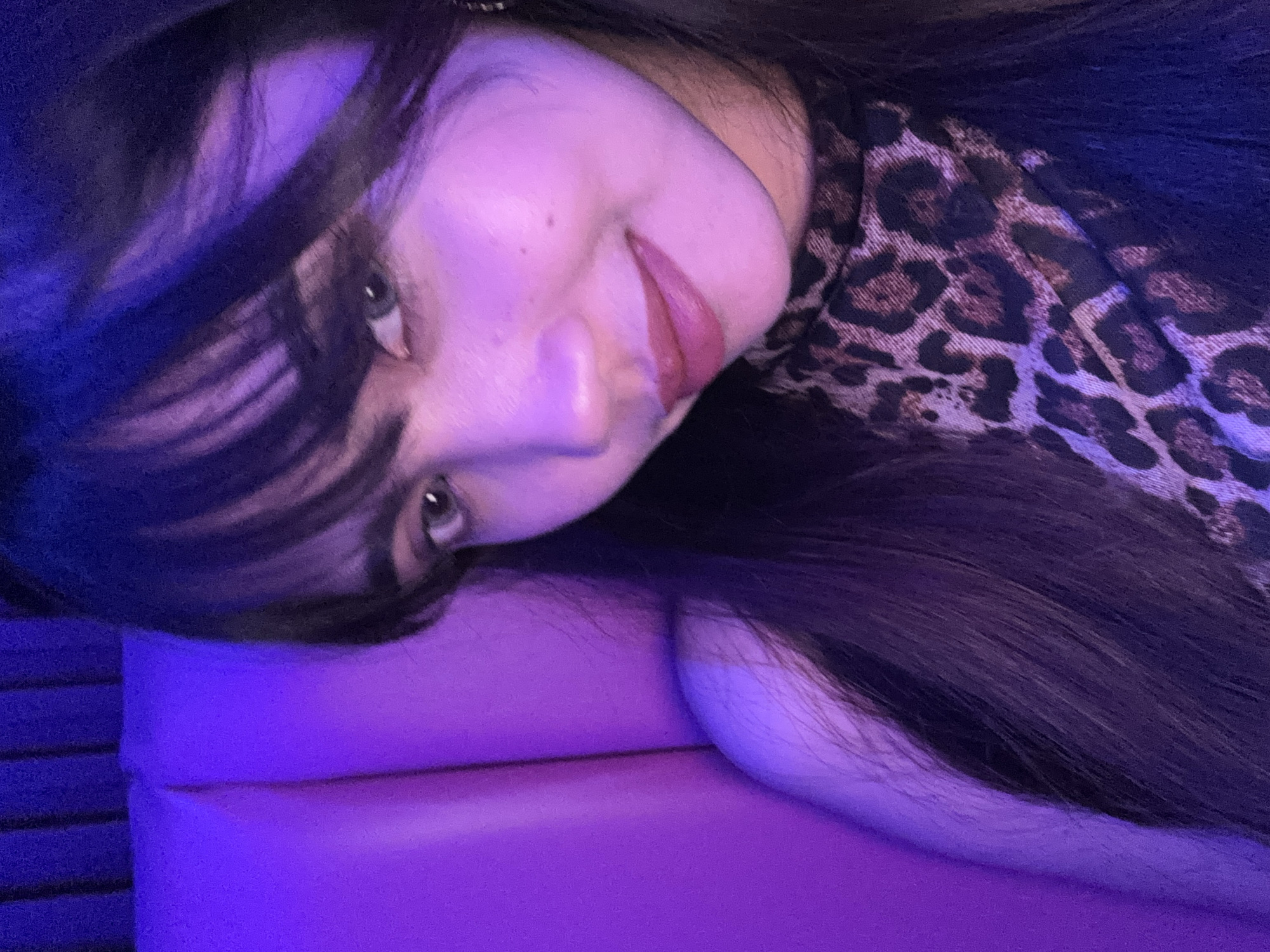 A young woman with dark hair and bangs is captured in a close-up, selfie-style photograph. She is wearing a leopard-print top. The background consists of a plush, textured surface bathed in purple and blue ambient lighting. The scene suggests an indoor setting, possibly a booth or seating area with a distinct purple hue dominating the environment. The woman is looking towards the camera with a subtle smile. The image quality is slightly grainy, with the lighting casting soft shadows and highlighting her facial features, particularly her eyes and lips, which appear to be adorned with makeup. There is no visible indication of the specific location within Davao City, Philippines.