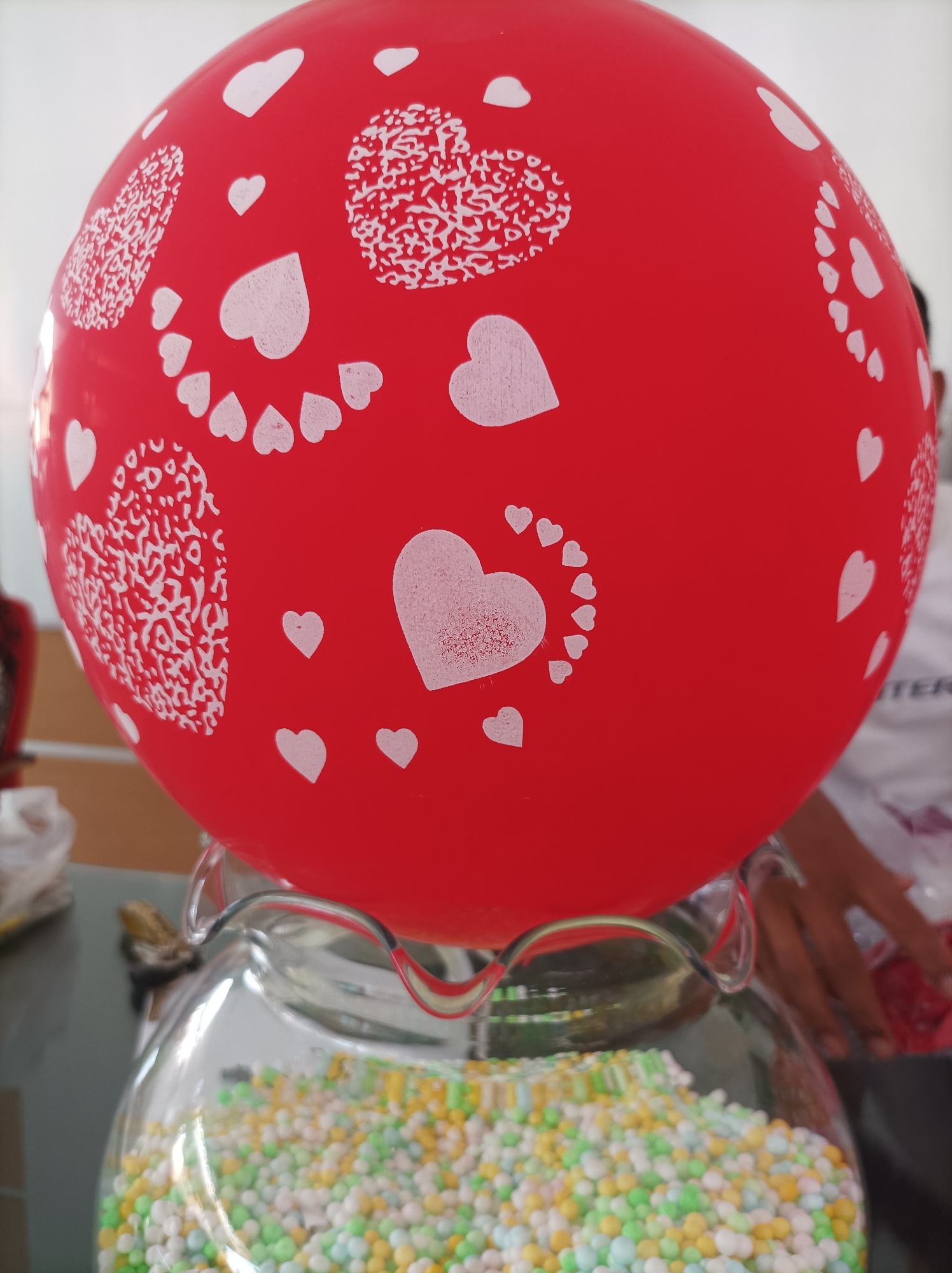 A red balloon with white heart designs is positioned above a clear glass jar filled with small, colorful spherical candies. The balloon is decorated with various sizes and styles of white hearts, some filled with a textured pattern and others solid. The glass jar has a wavy rim, and the candies inside are a mix of light blue, green, yellow, and white. In the background, a blurred image of hands and a person wearing a white t-shirt is visible, suggesting an indoor or outdoor setting with other people present. The environment appears to be a tabletop with other objects and a wall in the background. The scene is likely from a celebration or event in Ringinanom, Indonesia, possibly a party or a candy shop.