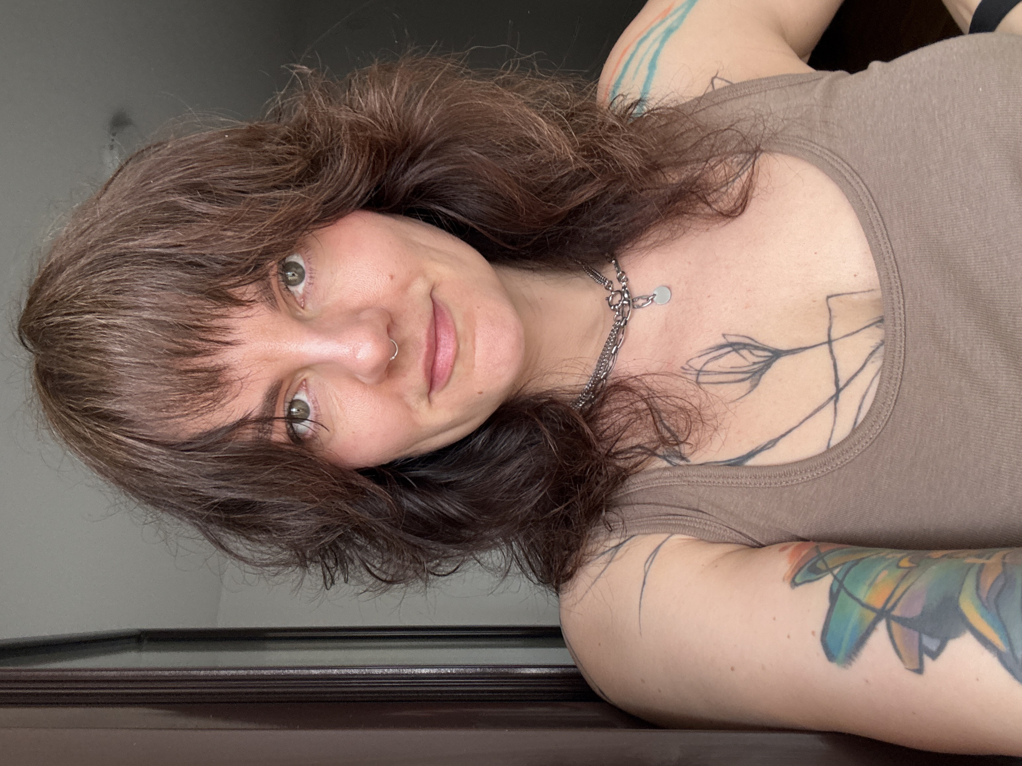A medium shot of a woman with brown hair and brown eyes, wearing a brown tank top and a silver chain necklace, positioned against a grey wall and a dark wooden surface. Tattoos are visible on her arms and chest. The photograph appears to be taken indoors. The location is not discernible from the provided image. There is no information related to the city of Boreti, Montenegro, discernible in the image.