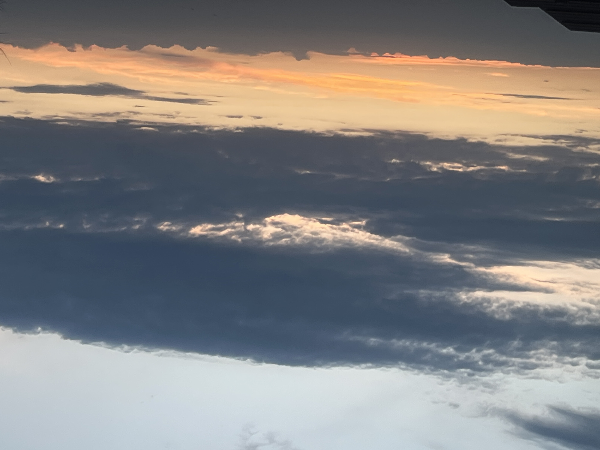 The image is a low-angle shot of a sky filled with layered clouds. At the top, there is a band of orange-tinted clouds indicating either sunset or sunrise. Below this, there are several layers of dark, gray clouds with patches of white where light is reflecting. The lower portion of the image shows lighter gray and white clouds, possibly lower altitude or thinner cloud cover. The sky appears overcast. There is no visible evidence to indicate the location is Votkinsk, Russia, or any people, subjects, objects, actions, or interactions within the scene.