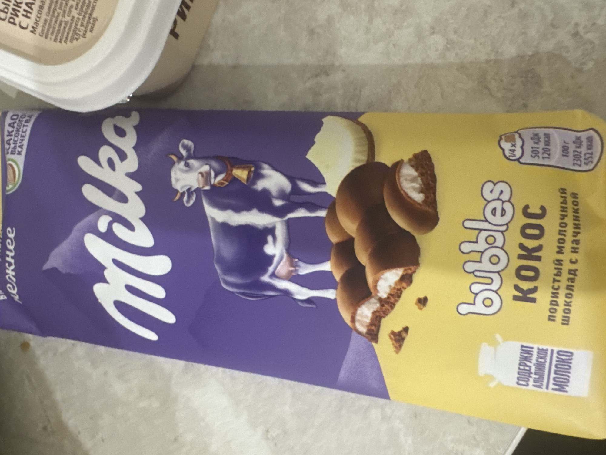 The image shows a package of Milka chocolate bars with coconut filling. The packaging is primarily purple and yellow, with a depiction of a cow and several chocolate bars. Text on the package is in Russian, indicating the product name, ingredients, and nutritional information. There is also a white container to the right of the chocolate bar package, likely another food product. The background appears to be a textured surface, possibly a table or countertop. There are no people or animals visible in the image. The scene does not provide any information about the specific location within the city of Saratov, Russia.