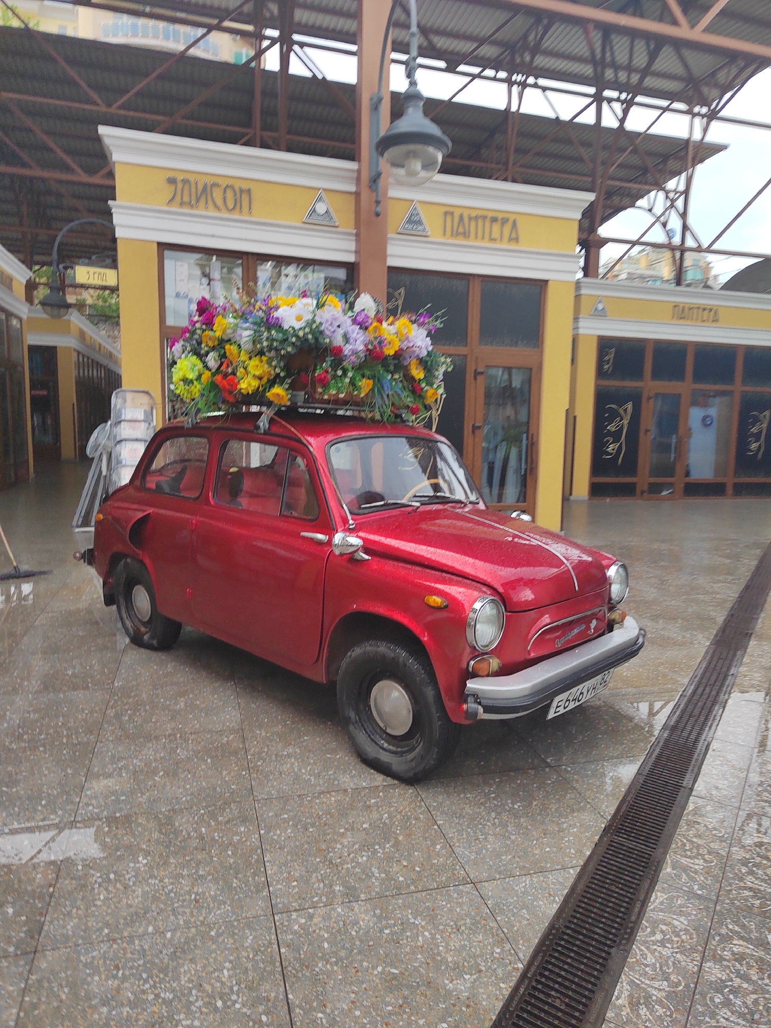 A small, red car is parked outdoors on a wet, tiled surface. The car has a large display of colorful flowers and greenery strapped to its roof. Behind the car, there are low, yellow buildings with the words "ЭДИСОН" and "ΠΑΠΤΕΓΑ" printed in yellow letters above the entrances. A tall, old-fashioned streetlight stands between the car and the buildings. The sky is overcast, suggesting the possibility of rain. The location is in the city of Unknown, Unknown.