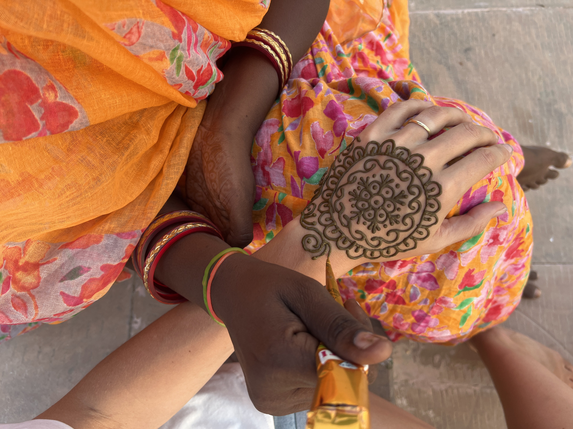 In Pushkar, India, a person is receiving a henna tattoo on their hand from another individual. The tattoo is a circular, intricate design on the back of the left hand. The artist, with darker skin, holds a cone applicator with their right hand and is in the process of drawing the design. The recipient, with lighter skin, has a delicate gold ring on their left ring finger. Both individuals are adorned with colorful clothing and jewelry. The person receiving the henna is wearing a vibrant orange and pink floral patterned garment, and the artist is wearing a bright orange fabric draped over their shoulder, with multiple bangles on their wrists. The background shows a paved surface, suggesting an outdoor setting.