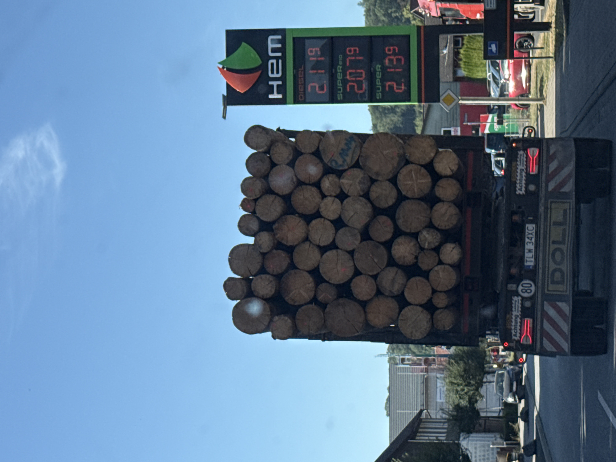 A lumber truck loaded with logs drives on a street in Walkenried, Germany. The truck is in the foreground, with a gas station sign to its right displaying prices for diesel and super gasoline. The sign shows "HEM" at the top, a logo, and prices "2.119" for diesel, "2.079" for SUPER E10, and "2.139" for SUPER. The truck's rear has a license plate reading "TLW 34XC" and "80" indicating a speed limit. Other vehicles are visible on the street, including a red car and a silver car in the background. Buildings and trees line the street, under a clear blue sky.