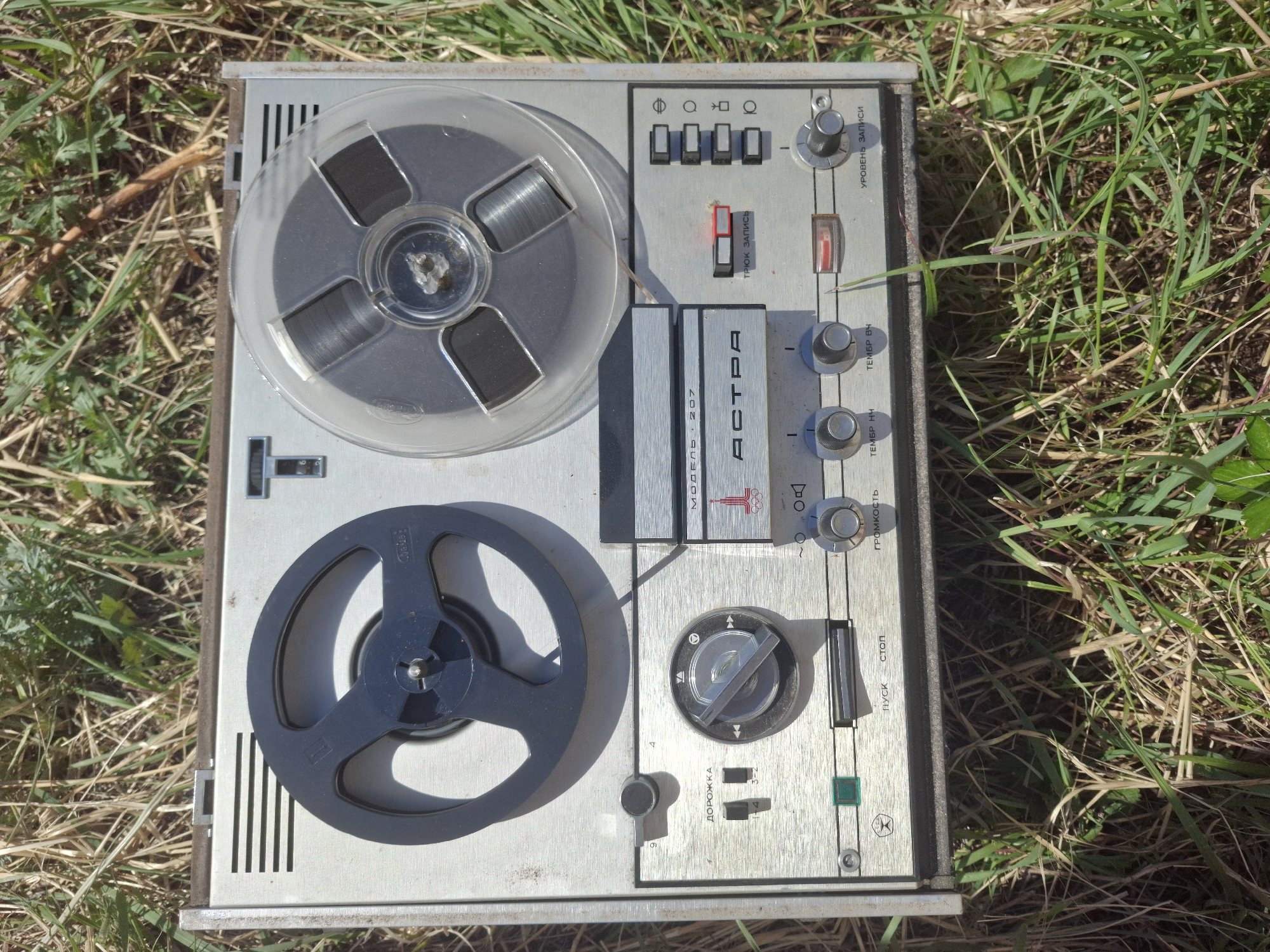 The image shows a vintage, silver-colored, reel-to-reel tape recorder sitting on a patch of grass. The recorder has two reels, one with clear plastic and the other with dark material. Various control buttons and knobs are visible on the right side of the recorder. The front panel includes the name "АСТРА" (ASTRA) and "МОДЕЛЬ 207" (MODEL 207). The text on the control panel is in Russian, indicating that the recorder was likely made in the Soviet Union or a country influenced by Soviet technology. The scene lacks identifying characteristics that would allow for pinpointing its location in Chaikovsky, Russia.