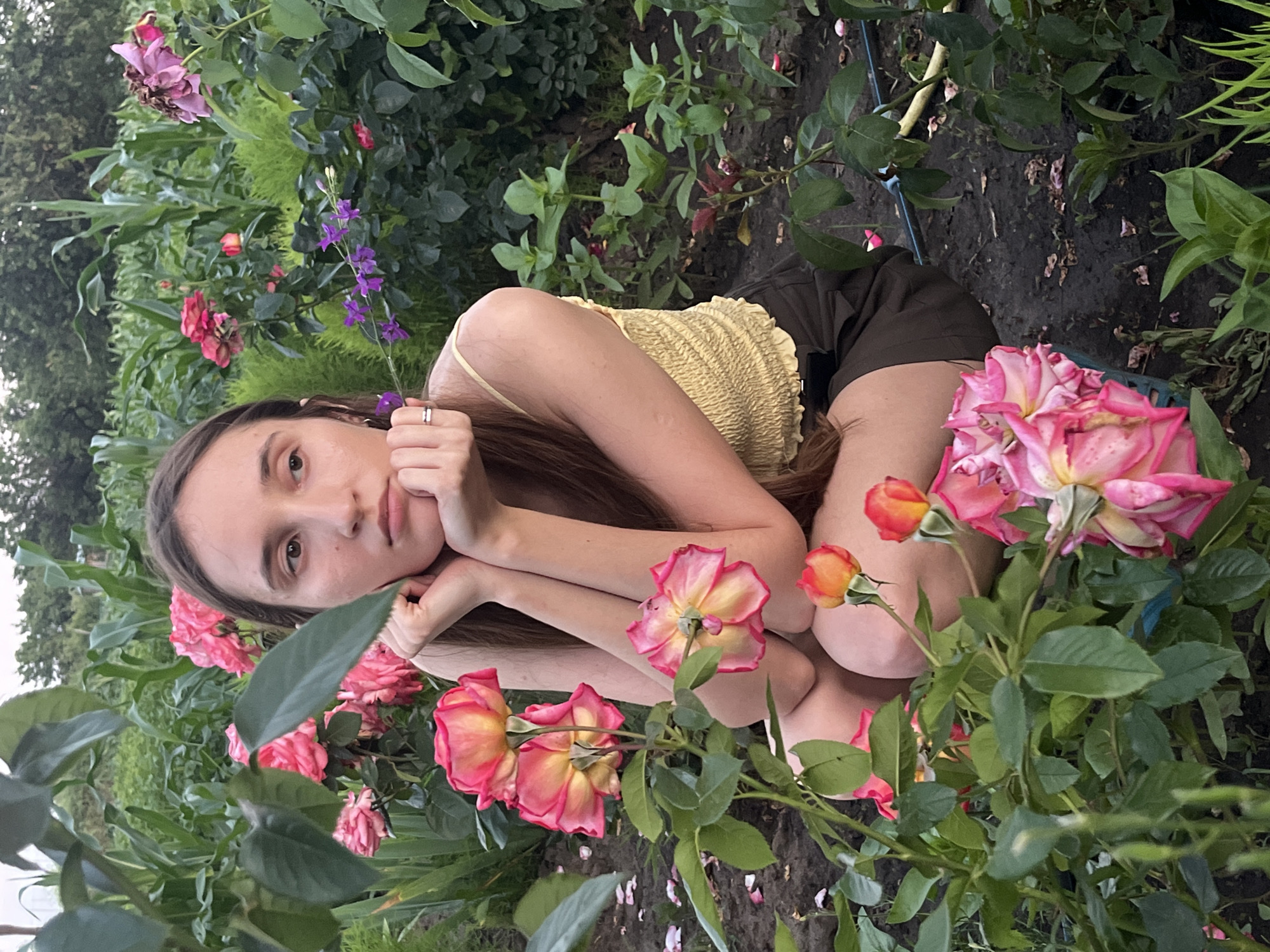 A young woman with long brown hair is partially visible in a garden. She is wearing a yellow, smocked crop top and dark shorts. Her arms are crossed in front of her, and she rests her head on her hands, looking towards the camera with a neutral expression. She has a ring on her left ring finger.
The setting is a lush garden filled with various plants and flowers. Prominent among them are several large, pink and yellow roses in bloom, some with hints of red. There are also smaller purple flowers on a delicate stem, and dense green foliage throughout the scene. The ground appears to be dark soil, with some fallen petals scattered around.
The lighting suggests it is daytime, possibly overcast or late afternoon, as the light is soft and diffused. The overall mood is serene and natural. There is no visible text in the image.