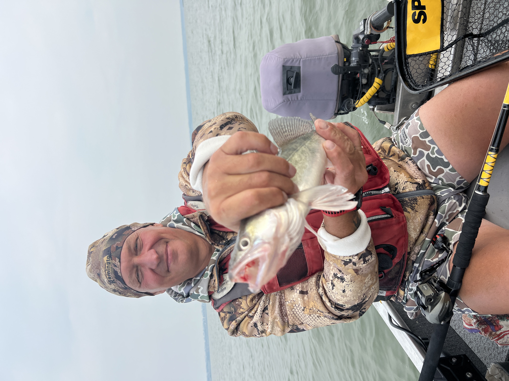 An outdoor medium shot captures a Caucasian man in a boat, holding a caught fish. He is wearing camouflage clothing, a red life vest, and a matching beanie. The man smiles at the camera, displaying the fish he is holding. The boat is on a body of water, with a cloudy sky overhead. An outboard motor is visible at the back of the boat. The man appears to be fishing in the city of Novopichugovo, Russia.