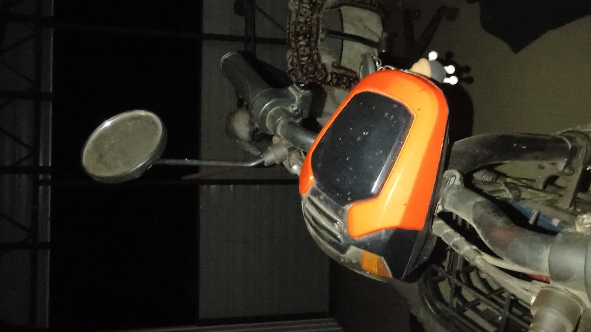 In a dimly lit, indoor or covered outdoor setting, the foreground of the image features the front of a motorcycle. The dominant colors are orange and black. The motorcycle's front fairing is a prominent orange and black structure, showing some dust. A black handlebar with a mirror attached to a silver stem is visible. A small, white, doll-like keychain with multiple white appendages hangs from the right side of the fairing. In the background, a dark, textured wall or partition is visible, with what appears to be some sort of shelving or structural beams above it. To the right, a patterned fabric and a chair are partially visible. The overall lighting is low, creating shadows and limited visibility of the surroundings. The scene is static, with no apparent action.