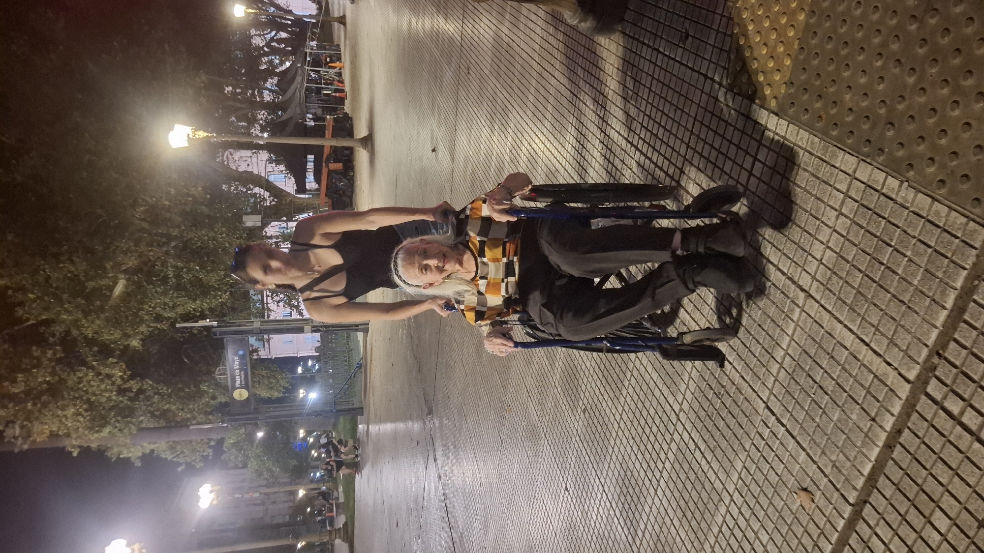 The image shows two people on a paved surface, likely a sidewalk, in an urban environment. An elderly woman with white hair and wearing a patterned shirt is seated in a blue wheelchair. A younger woman in a black top is pushing the wheelchair. The background features streetlights, trees, and buildings, suggesting a city setting. The area is illuminated with streetlights, indicating it is evening or night. A sign partially visible in the background reads "Plaza de Mayo" likely identifying this location in Buenos Aires, Argentina.