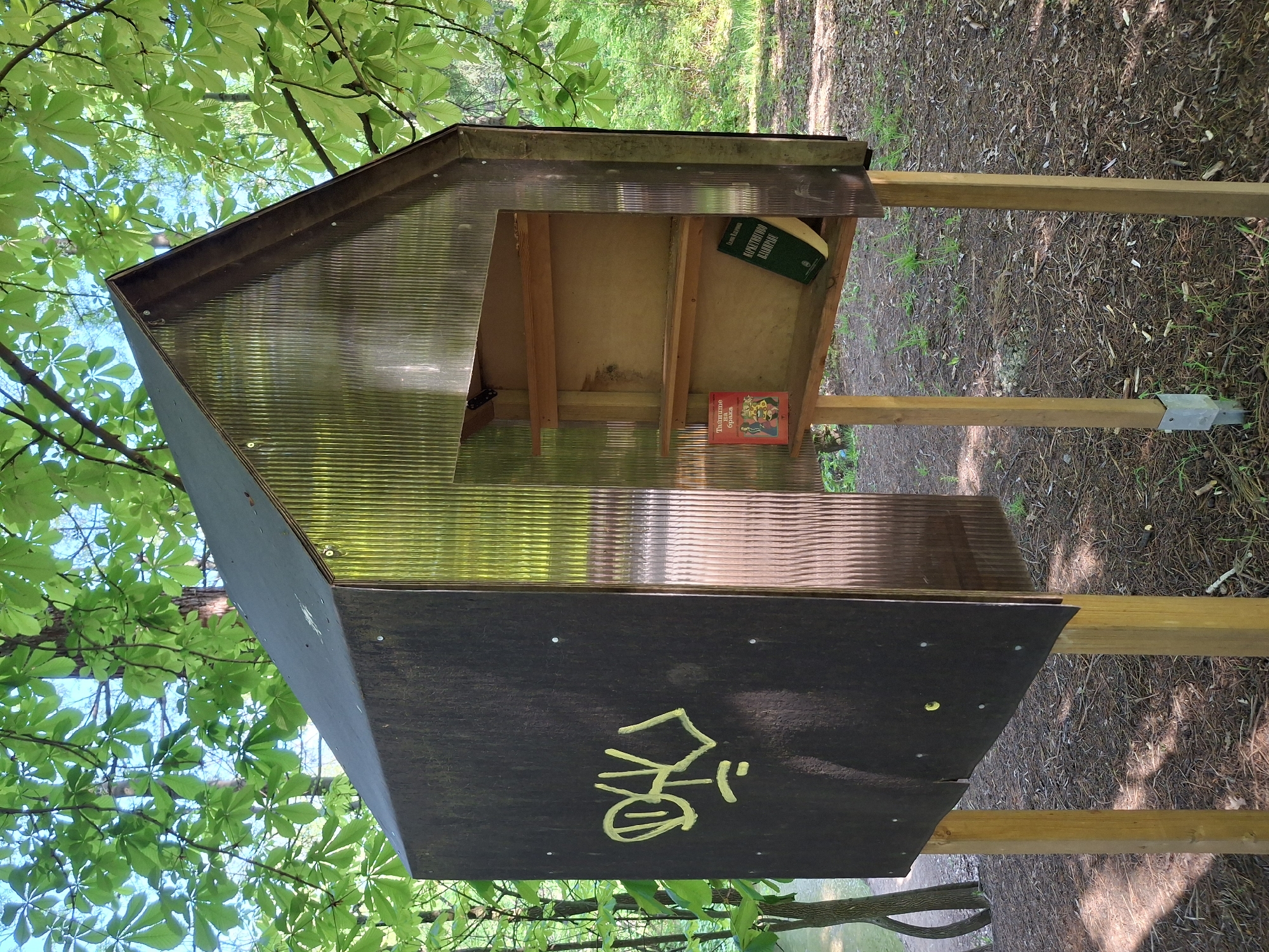 A tilted outdoor photograph captures a little free library with a small collection of books, situated in a wooded area possibly in Unknown, Unknown. The library structure features a black, slanted front with a yellow ideogram painted on it, likely a Chinese character. Its roof and sides are made of translucent corrugated plastic, revealing the shelf inside with two books visible: one with a green cover and the other with a red cover. The structure is supported by wooden posts embedded in the ground. The surrounding environment is characterized by fallen leaves, grass, and the foliage of trees, with sunlight filtering through the leaves creating a dappled lighting effect.